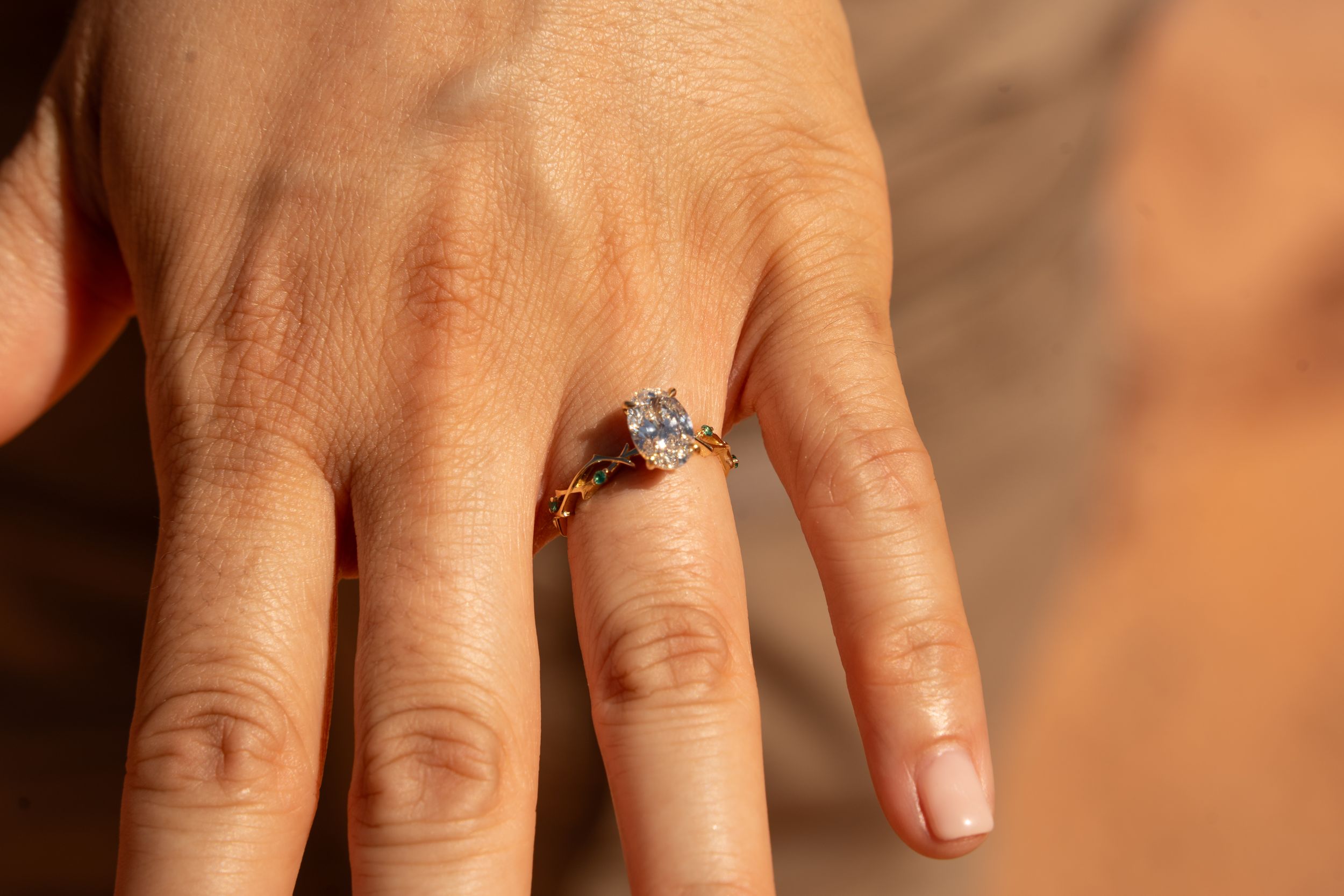 Close-up of a hand wearing a gold engagement ring with a large oval diamond solitaire and small green accent stones.