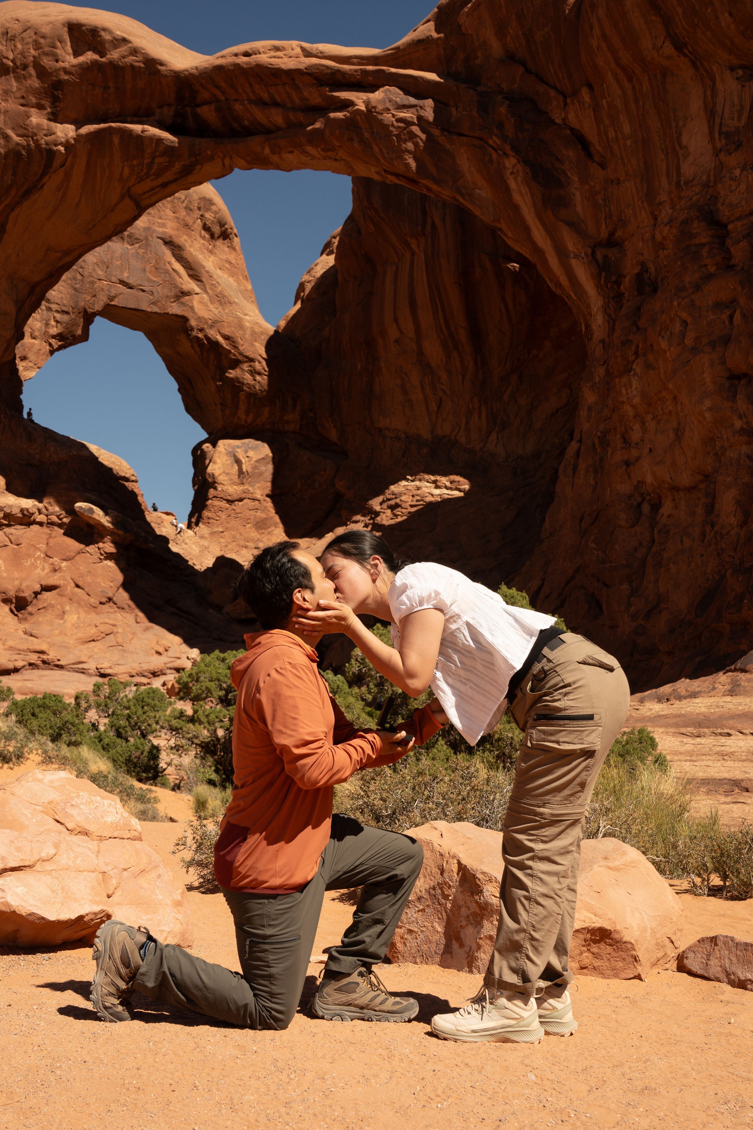 Man proposes to woman as she kisses him on one knee at Double Arch in Arches National Park, Utah desert landscape.
