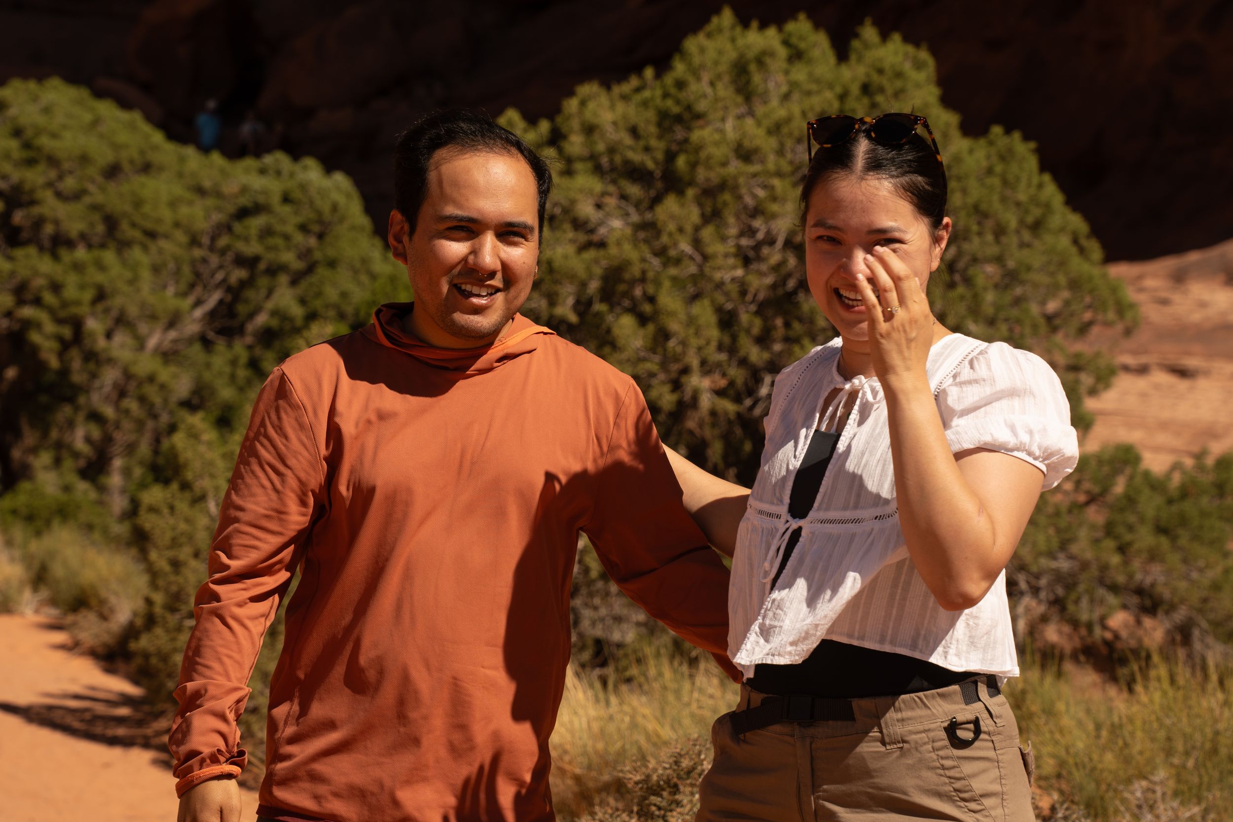 A man in an orange shirt and woman in white top smile together on a sunny red rock desert hiking trail.