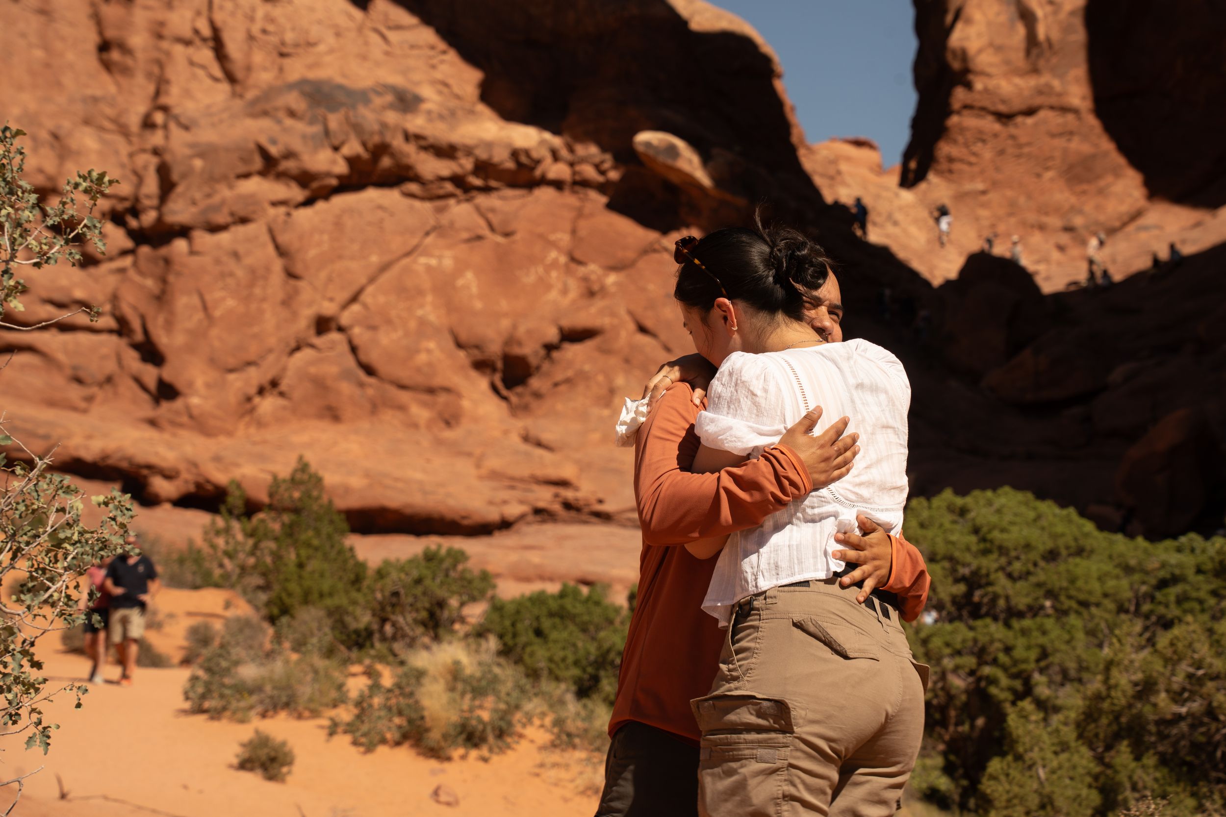 Couple embracing romantically among red rock formations in a desert landscape on a sunny day.