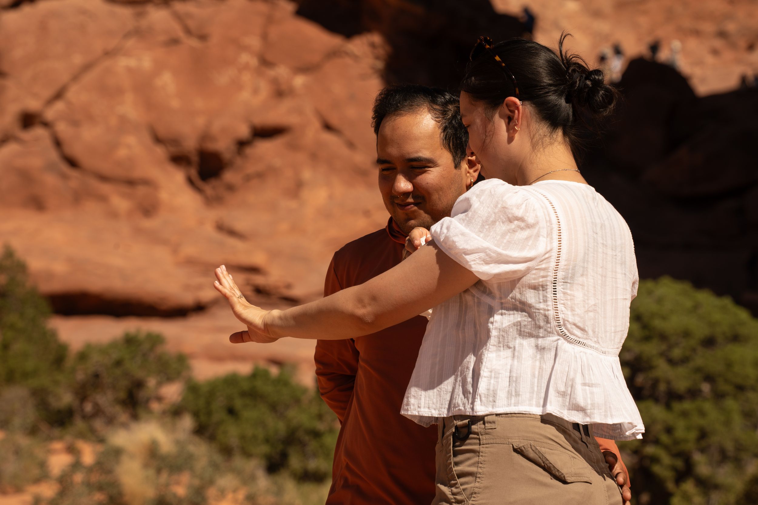Two people stand near red rock formations, and admiring her new engagement ring