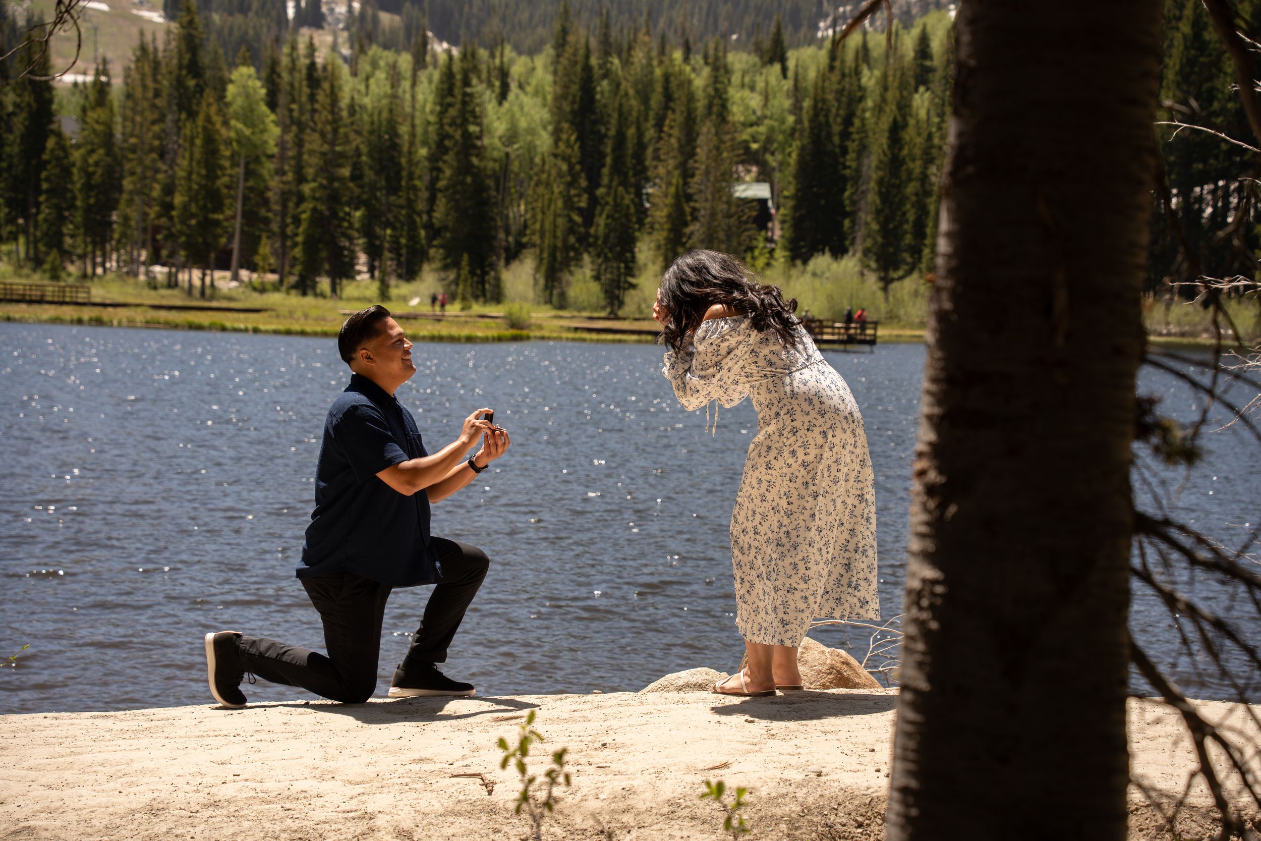 Man proposes on one knee to woman in floral dress by silver lake surrounded by pine trees in big cottonwood canyon