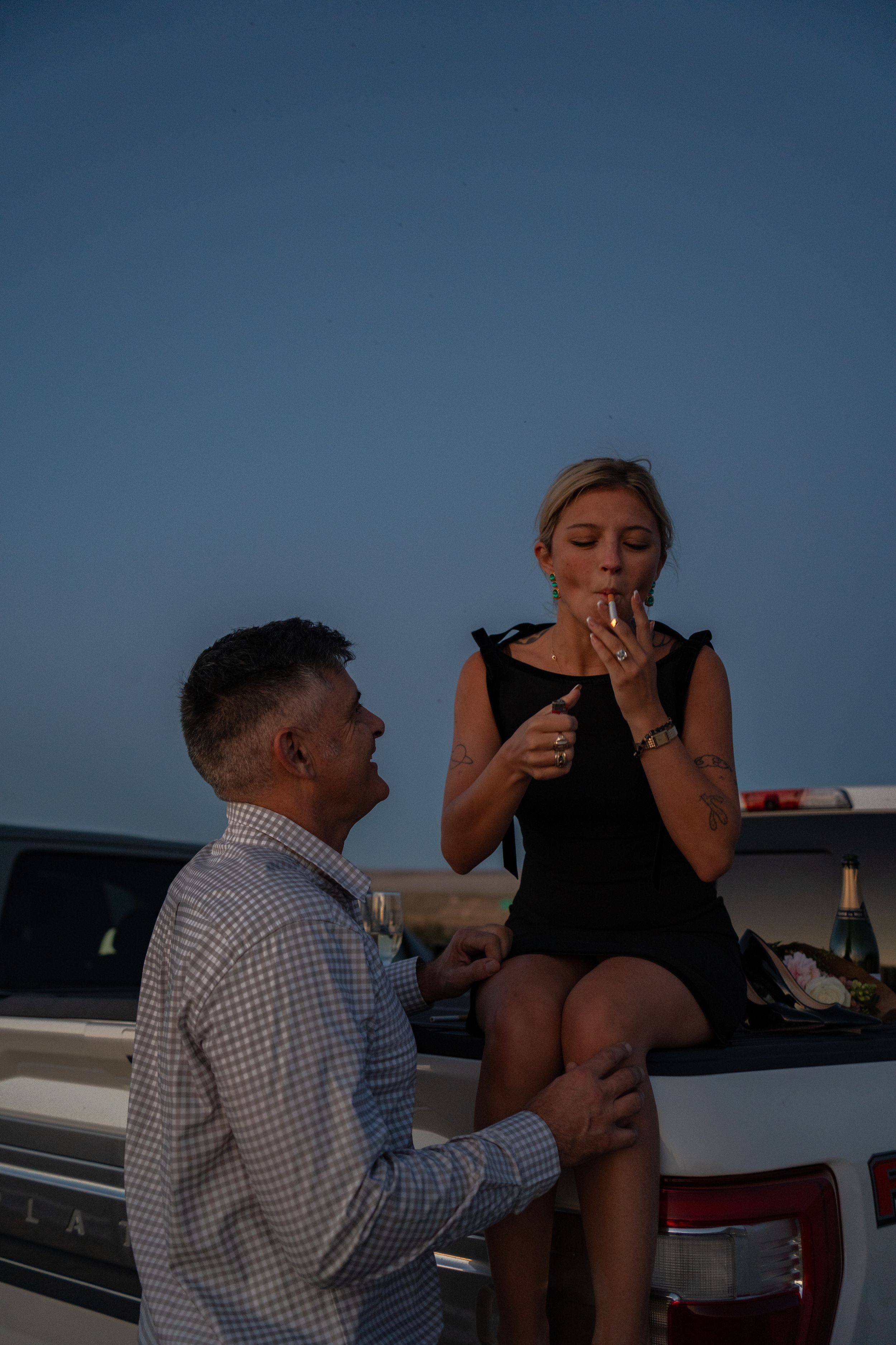 Man kneeling to propose to woman sitting on white truck tailgate at dusk, romantic outdoor engagement moment.