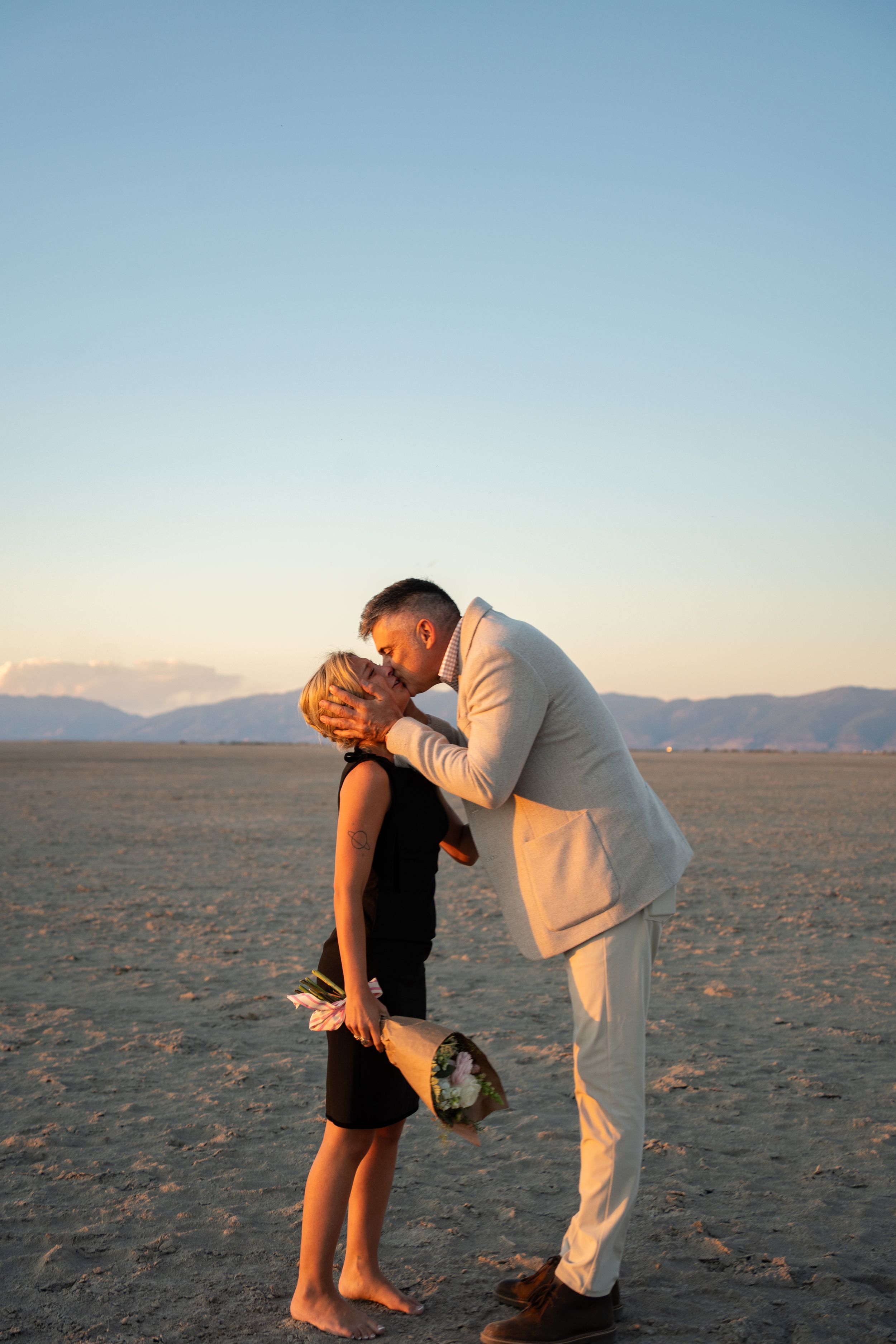 a newly engaged couple kissing on the salt flats of the great saltair holding flowers and celebrating a proposal
