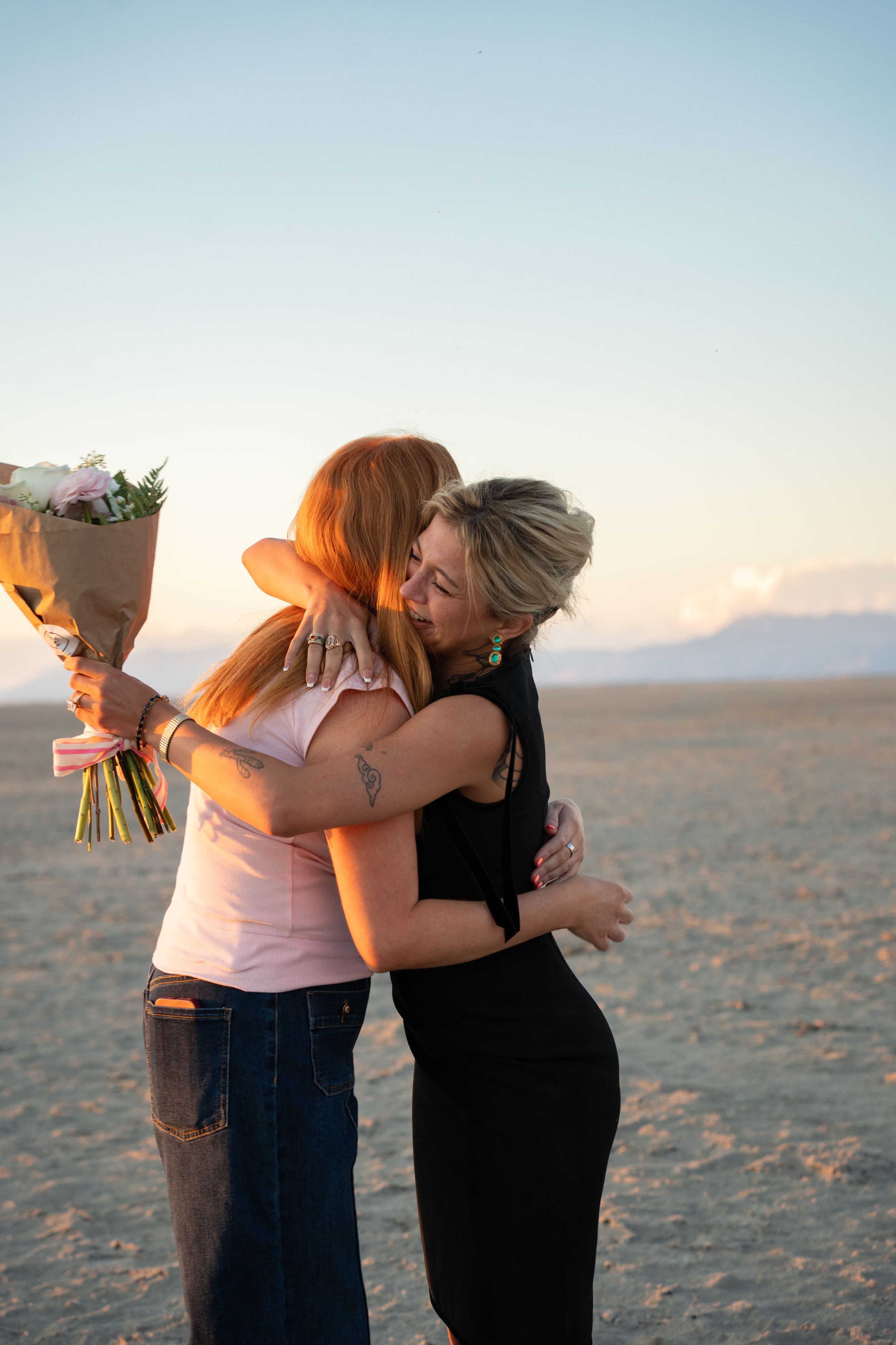 Two women embracing on salt frats at sunset, one holding a bouquet of flowers, sharing an emotional moment.