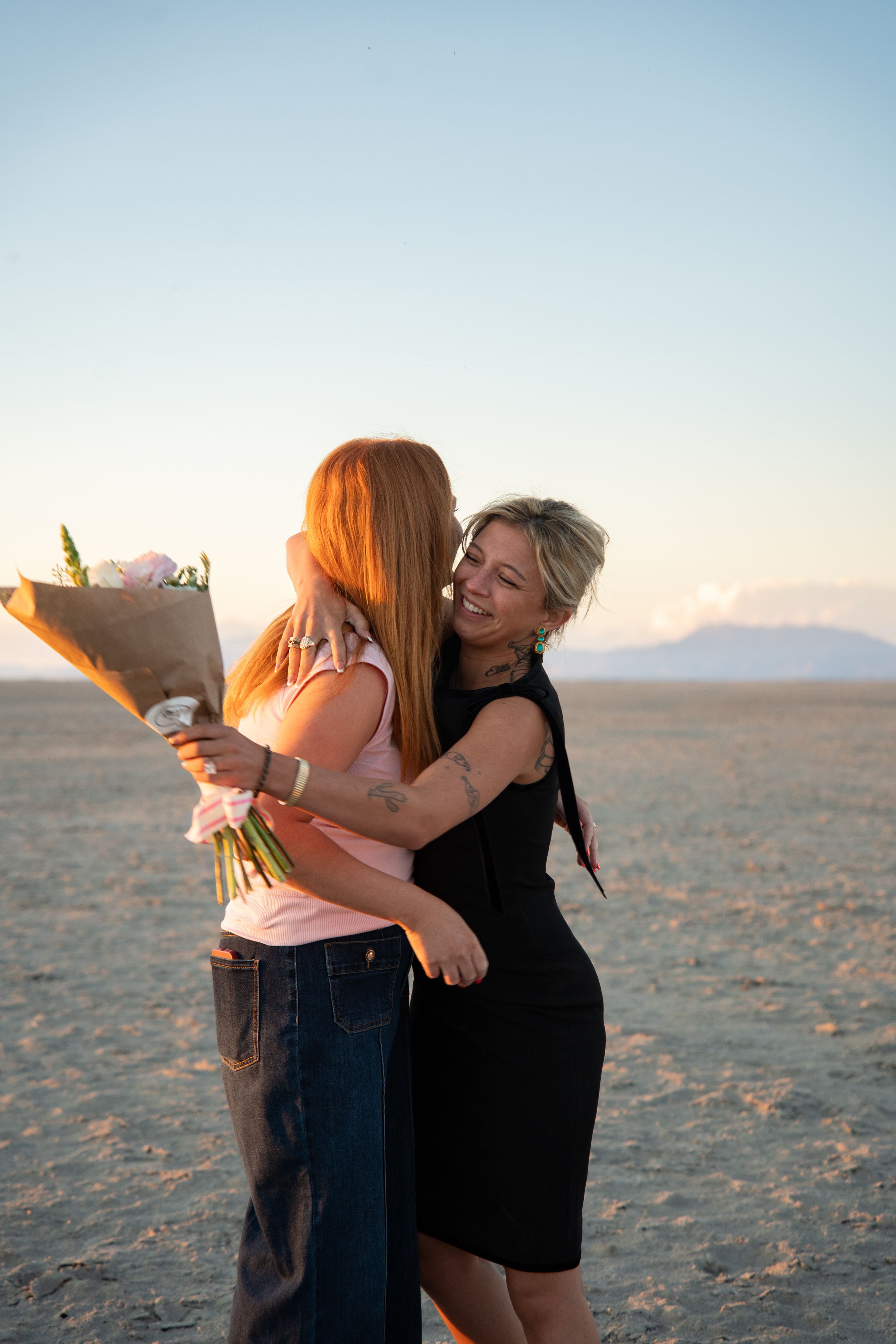 Two women hugging on salt flats at sunset, one holding a bouquet of flowers, smiling joyfully.