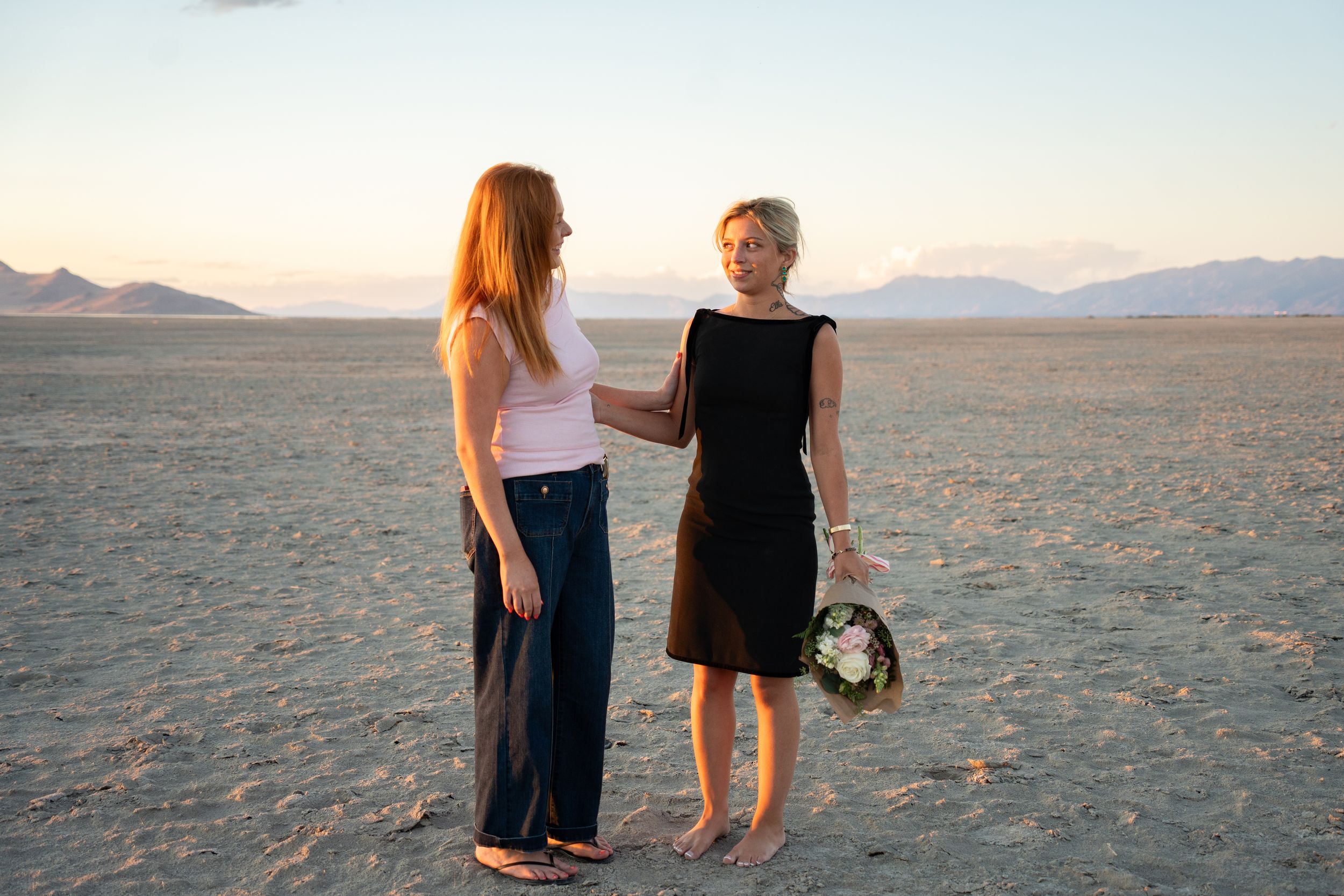 Two women standing on a salt flats of great saltair at sunset with rocky mountains in the background, one holding flowers.