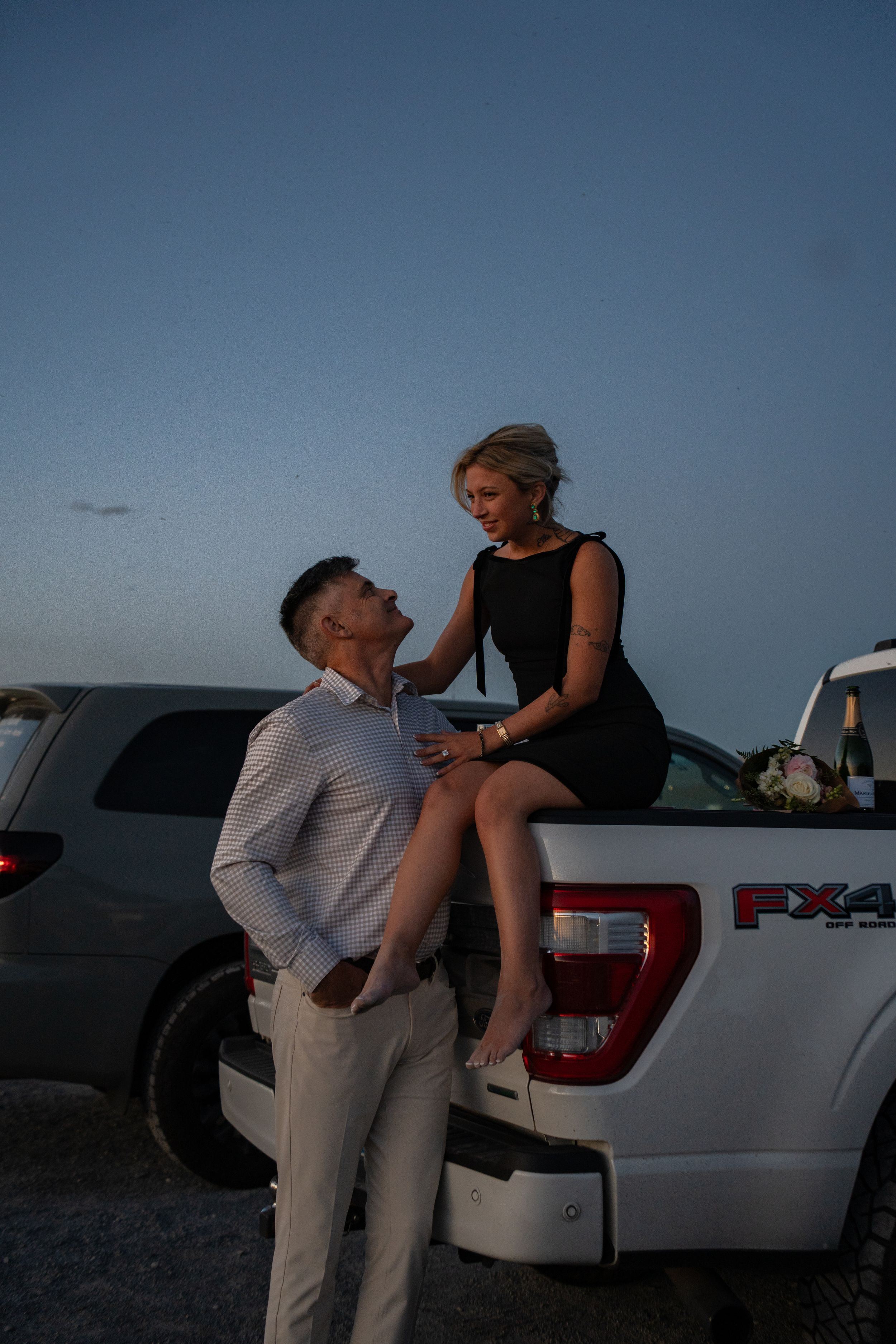 Couple at dusk, woman in black dress sitting on Ford FX4 truck tailgate, man in white shirt standing close at great saltair
