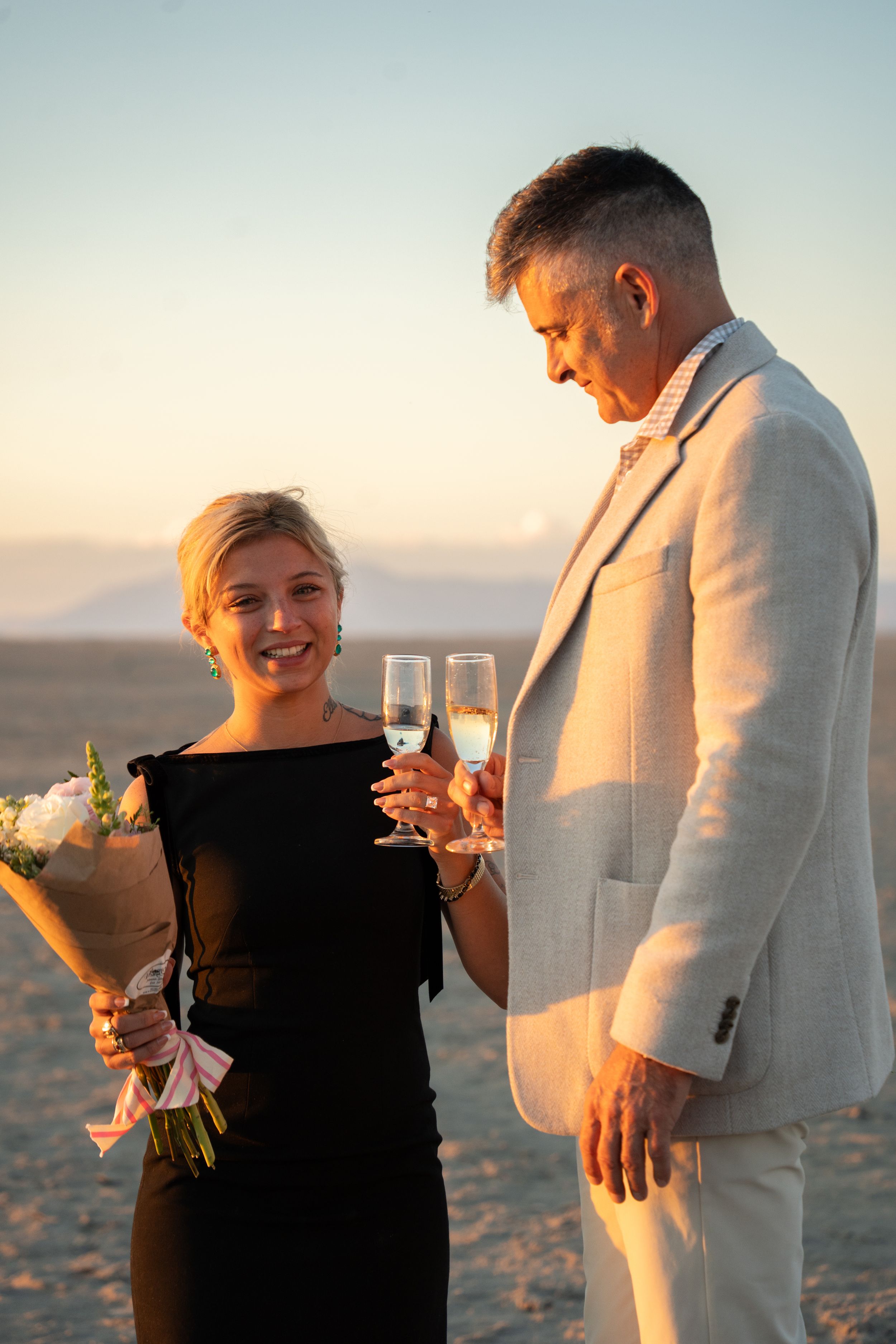 couple holding champagne glasses and flowers after proposal in the salt flats