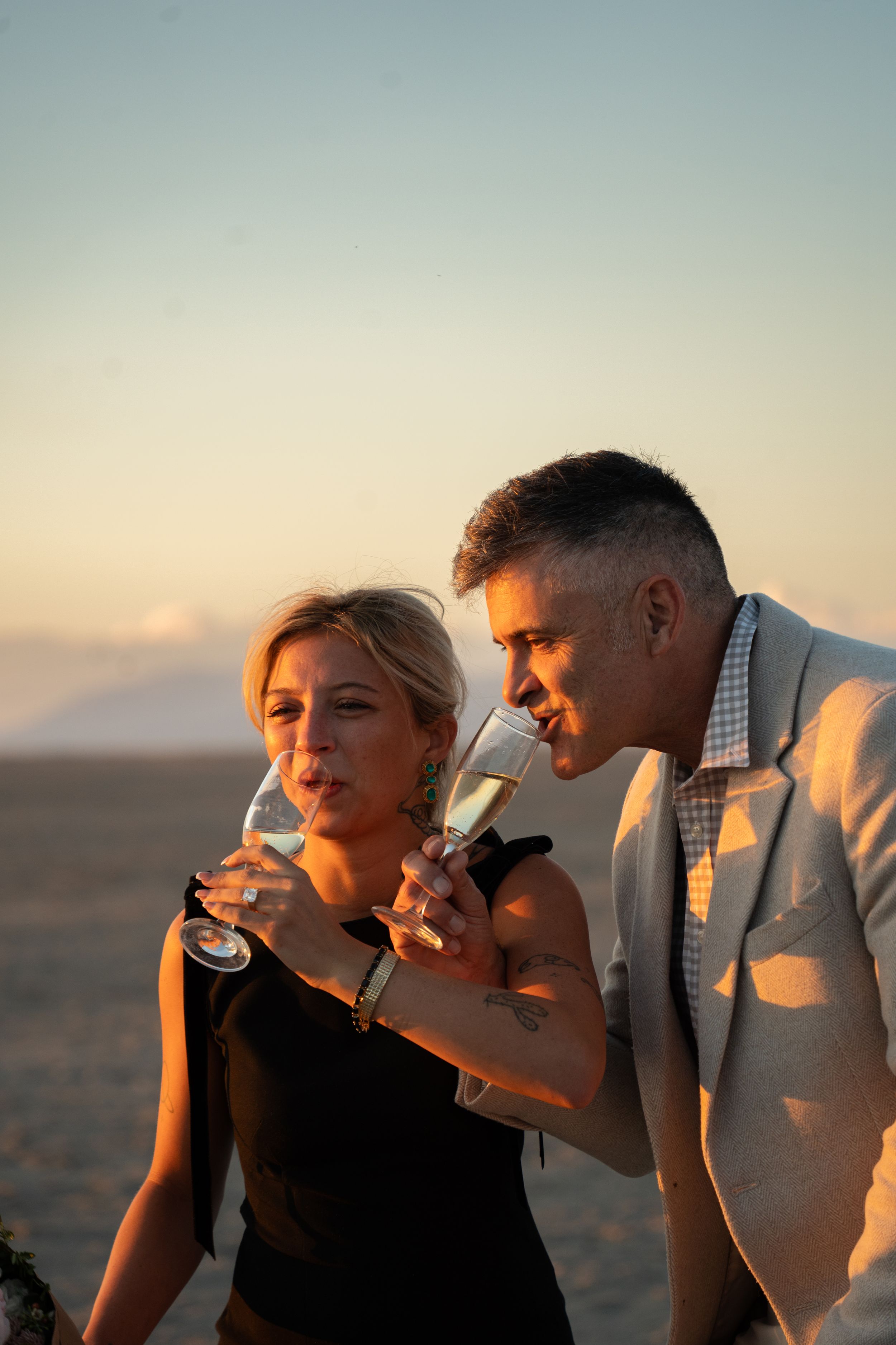 Elegant couple drinking champagne at sunset on salt flats, dressed in formal attire for a proposal celebration