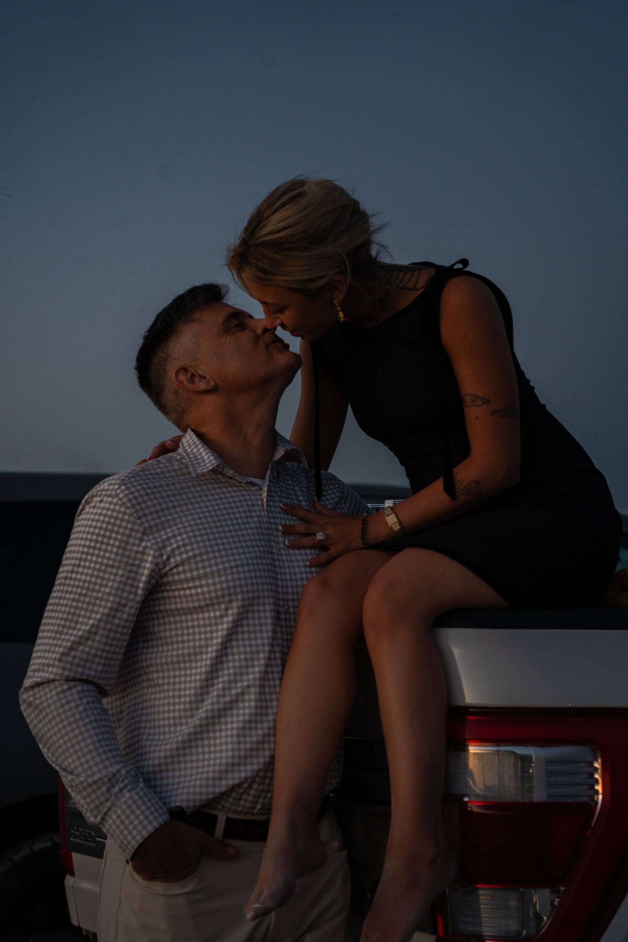 Romantic couple sharing a kiss at dusk, woman in black dress sitting on red truck, man in checkered shirt.