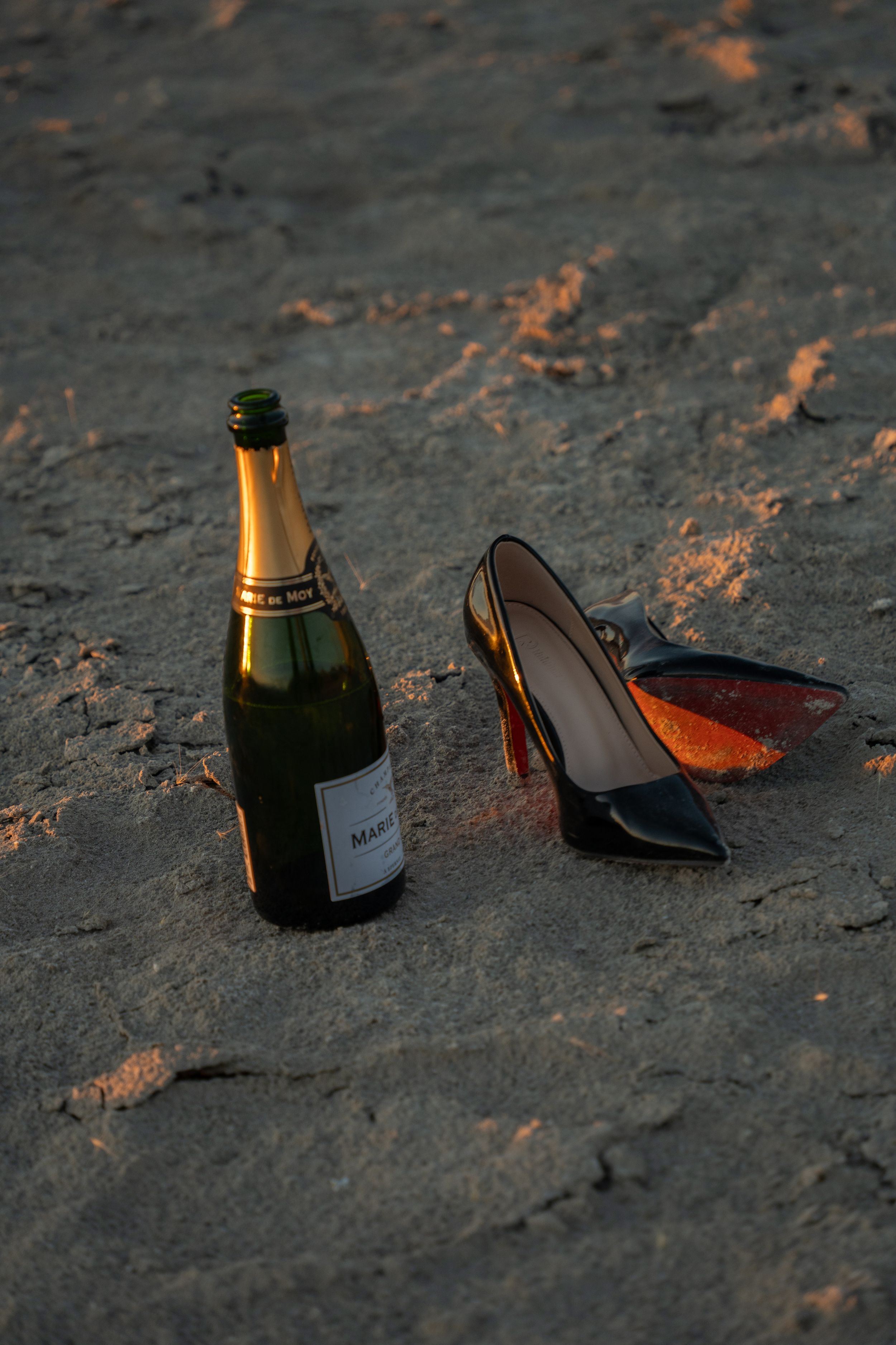 Champagne bottle and black high heel shoes with red soles resting on salt flat at golden hour sunset.