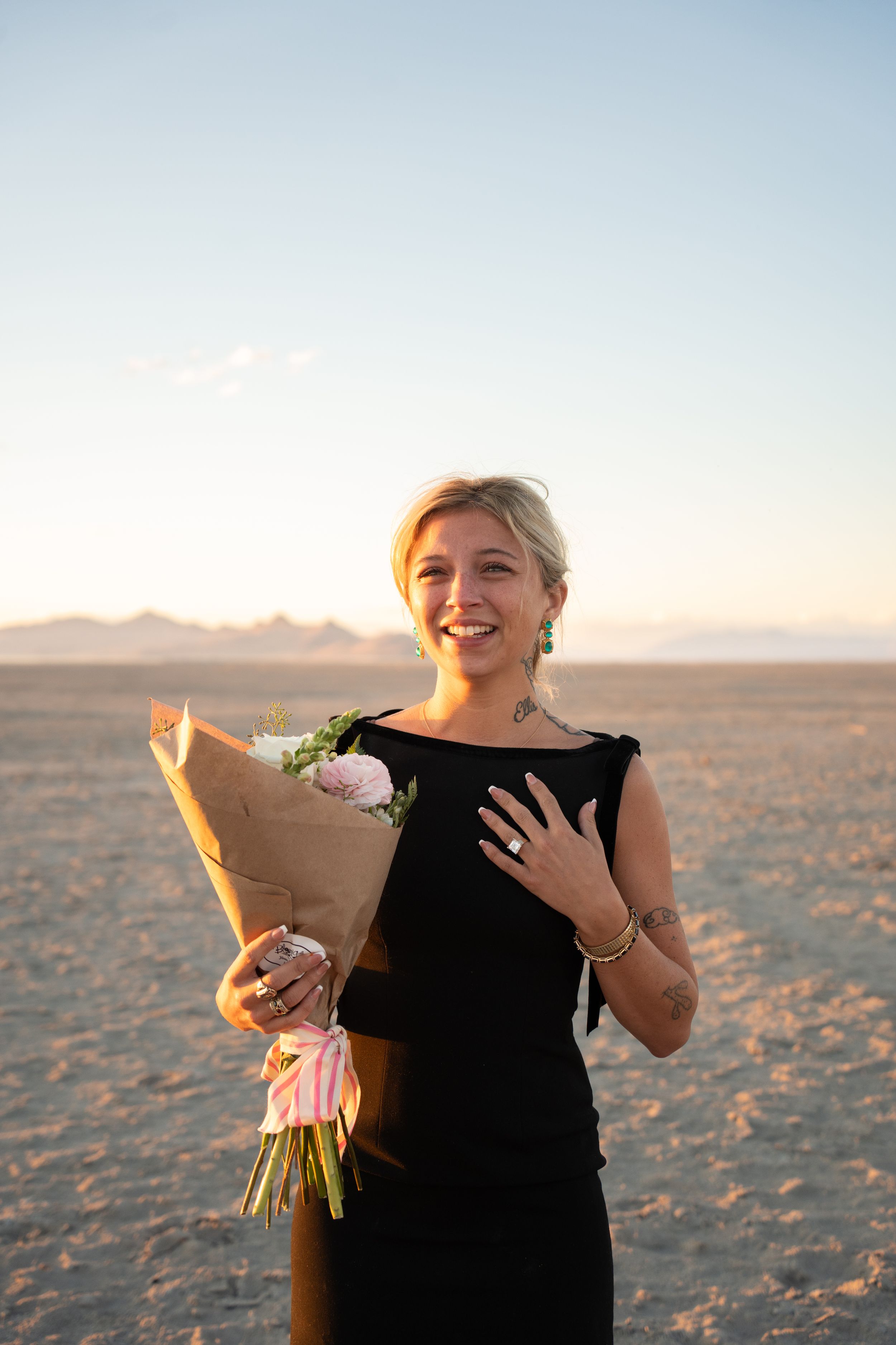 Smiling blonde woman in black dress holding flower bouquet gasping over her proposal displaying her new engagement ring
