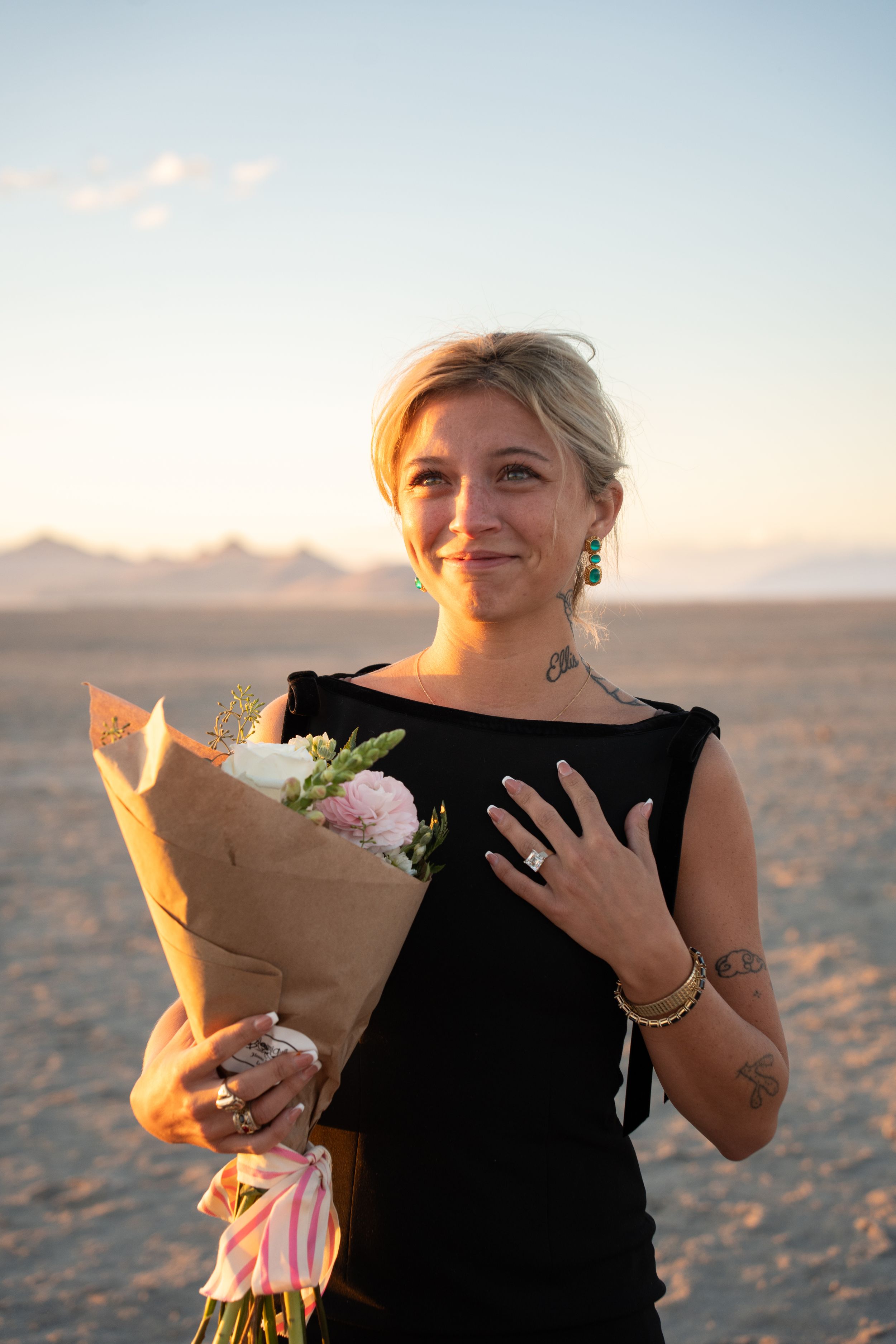 Smiling blonde woman in black dress holding flower bouquet on sandy flat at sunset, showing engagement ring.