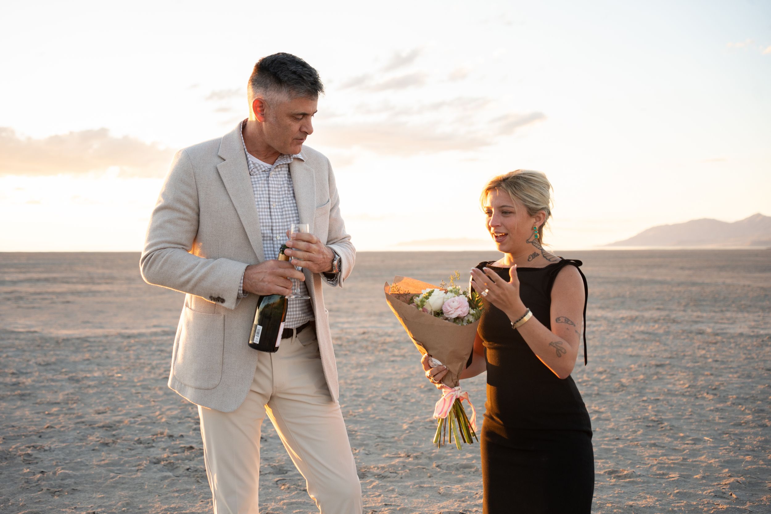 engaged couple celebrating on salt flats at sunset, man pouring champagne while woman holds a bouquet of flowers.