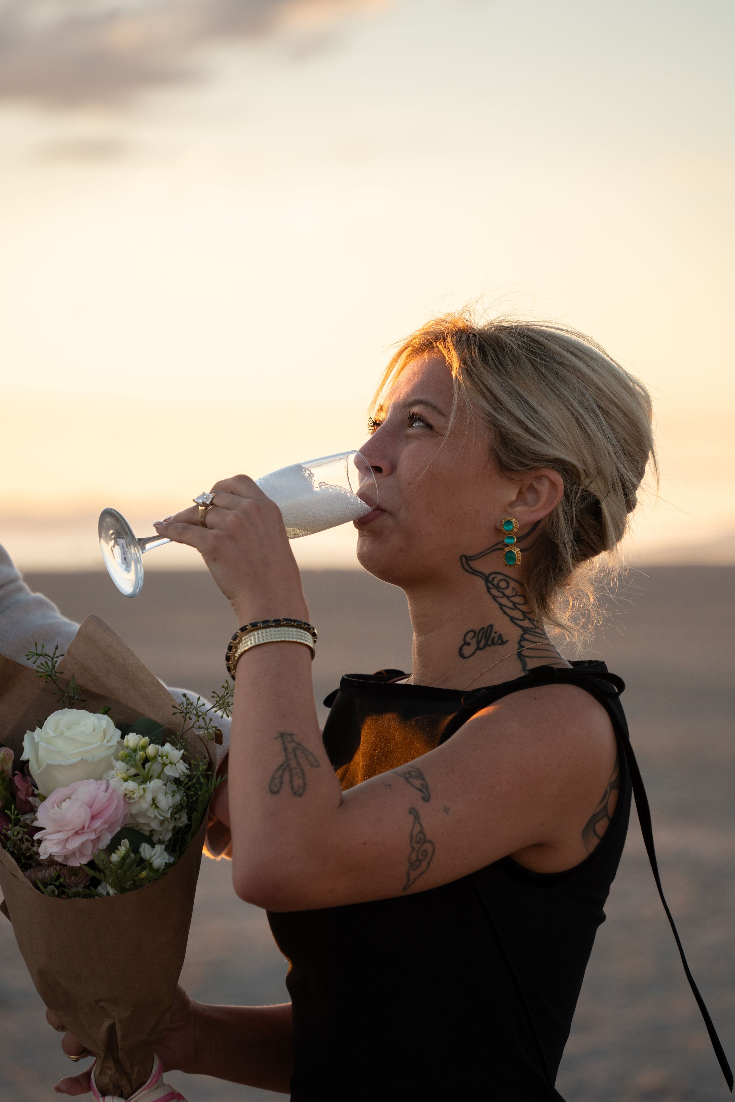 Tattooed blonde woman drinking champagne at sunset, wearing black dress and green earrings, holding a bouquet of flowers.