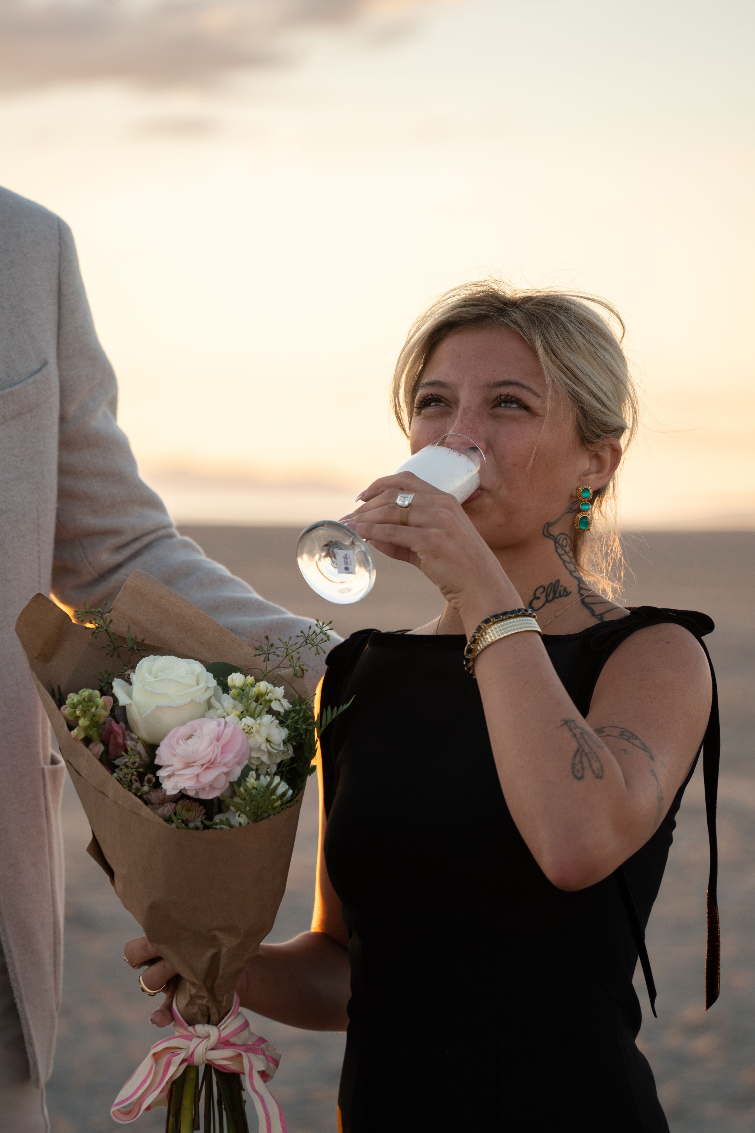 Woman in black dress drinks champagne holding flowers at sunset beach, tattoos visible, smiling joyfully and showing off her engagement ring