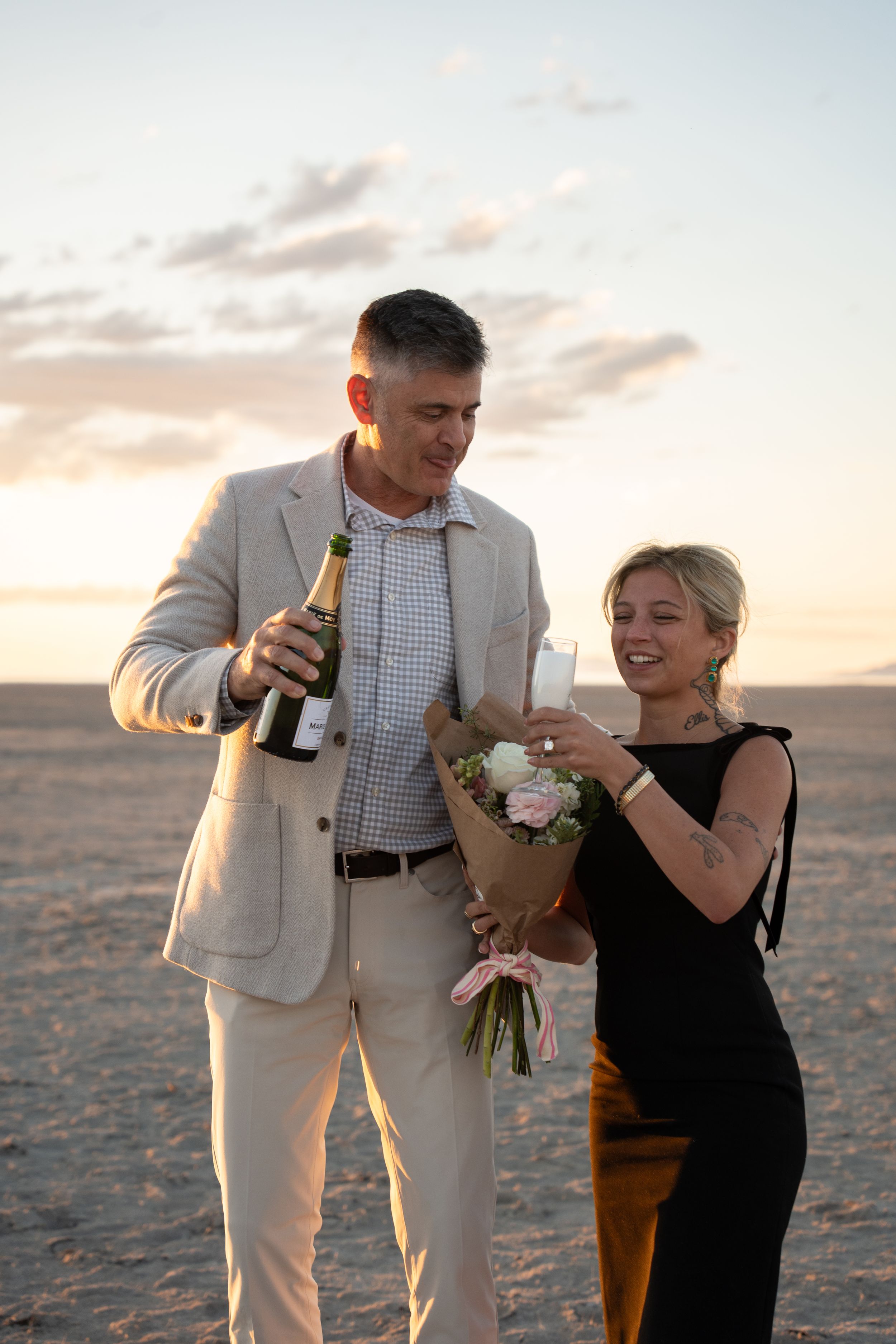 Couple celebrating on salt flats at sunset, man pouring champagne, woman holding bouquet of flowers, wearing elegant attire.