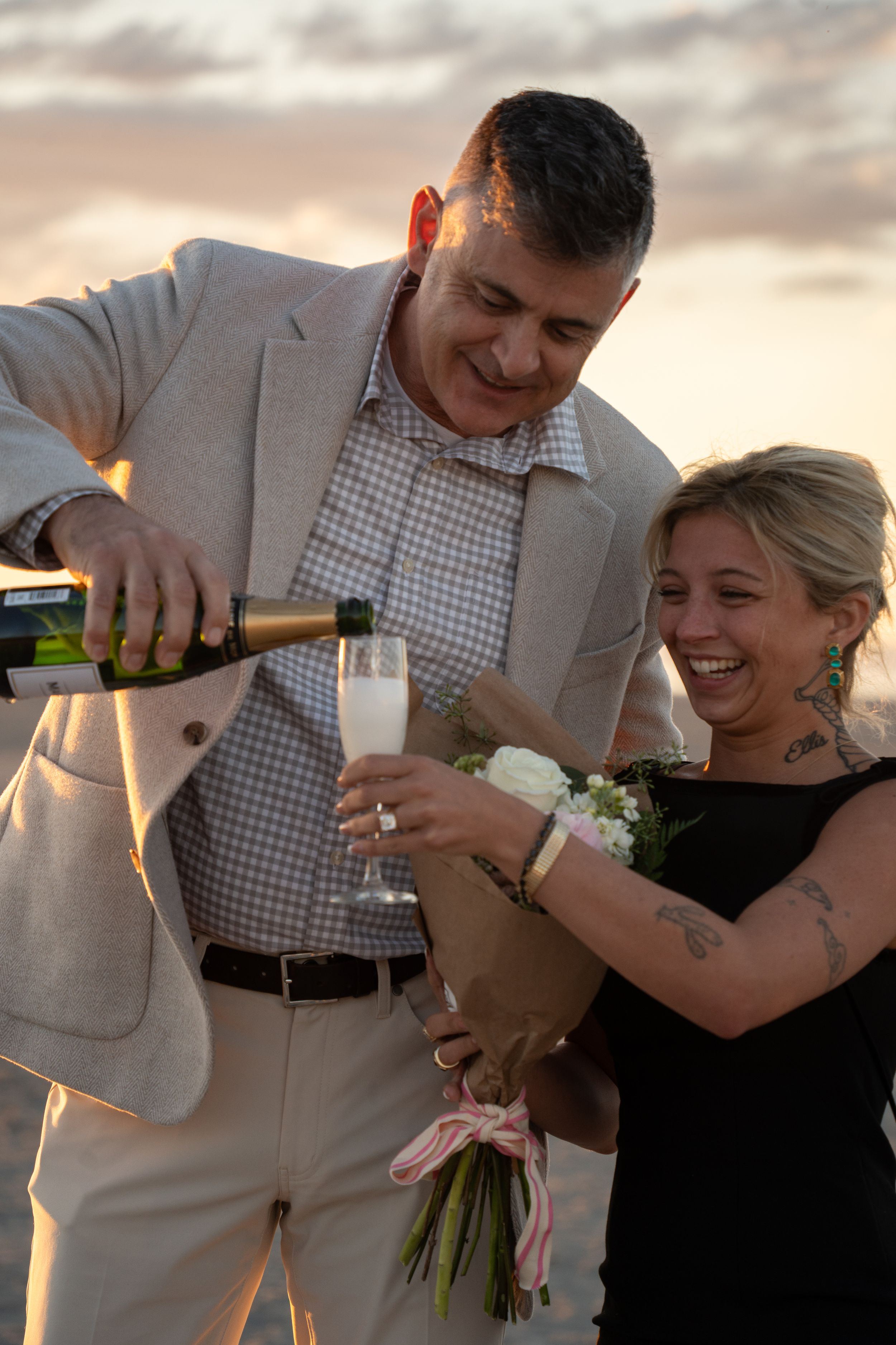 Man in beige suit pouring champagne for smiling tattooed woman holding white flower bouquet at sunset celebration of their proposal