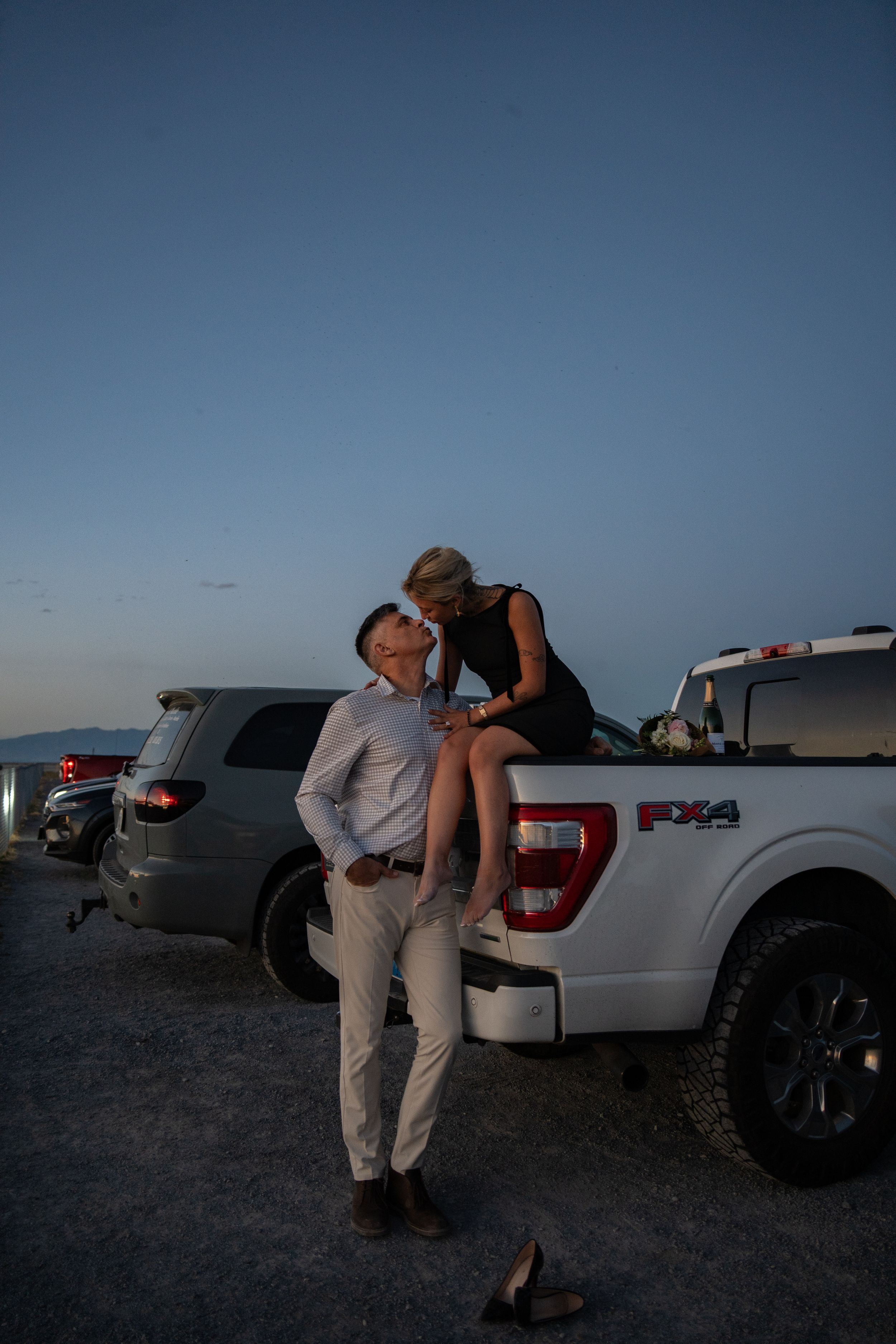 engaged couple sharing a romantic moment at dusk, woman sitting on white Ford F-150 truck bed leaning toward man standing beside it.