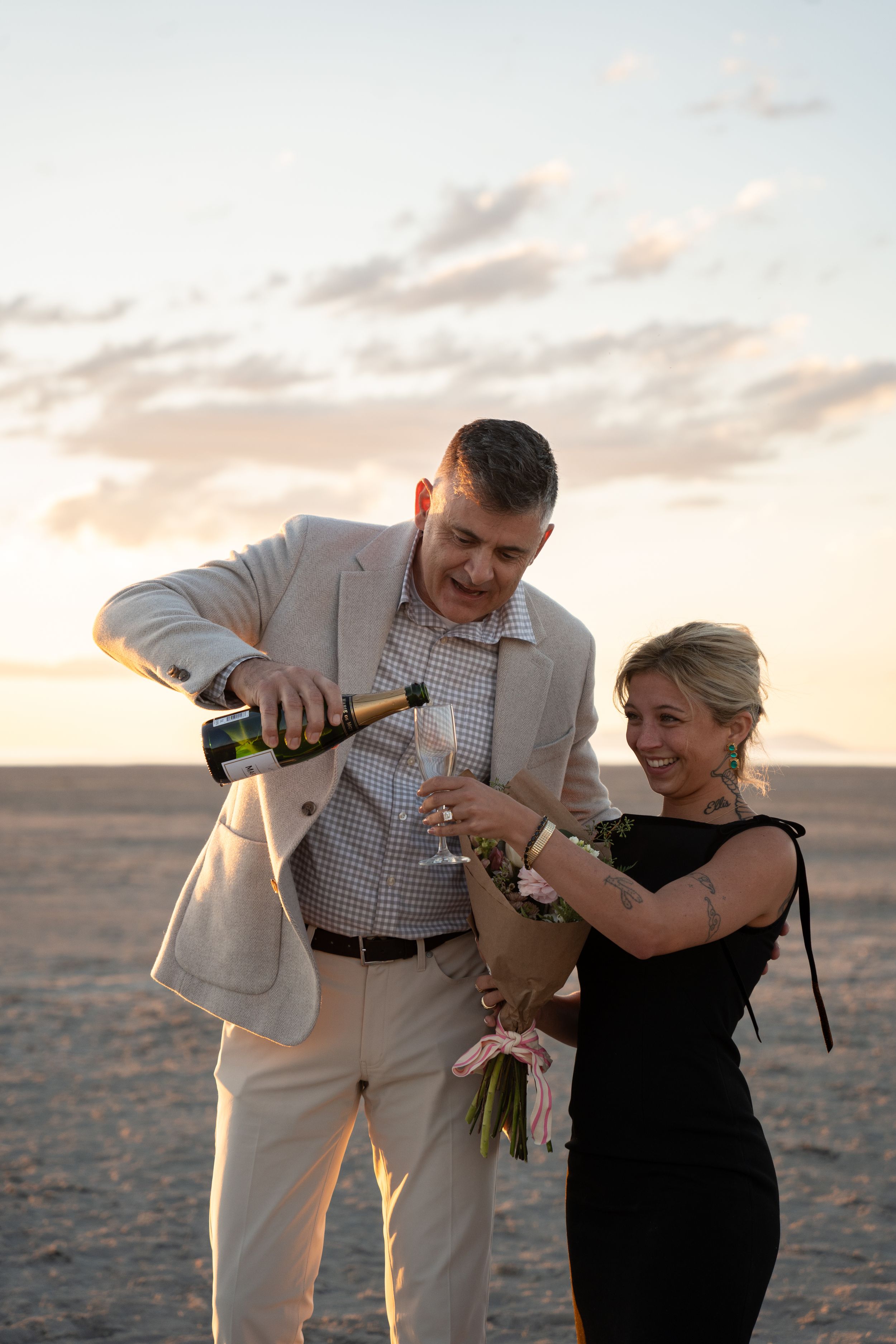 Couple celebrating on salt flat at sunset, man pouring champagne while woman holds bouquet of flowers.