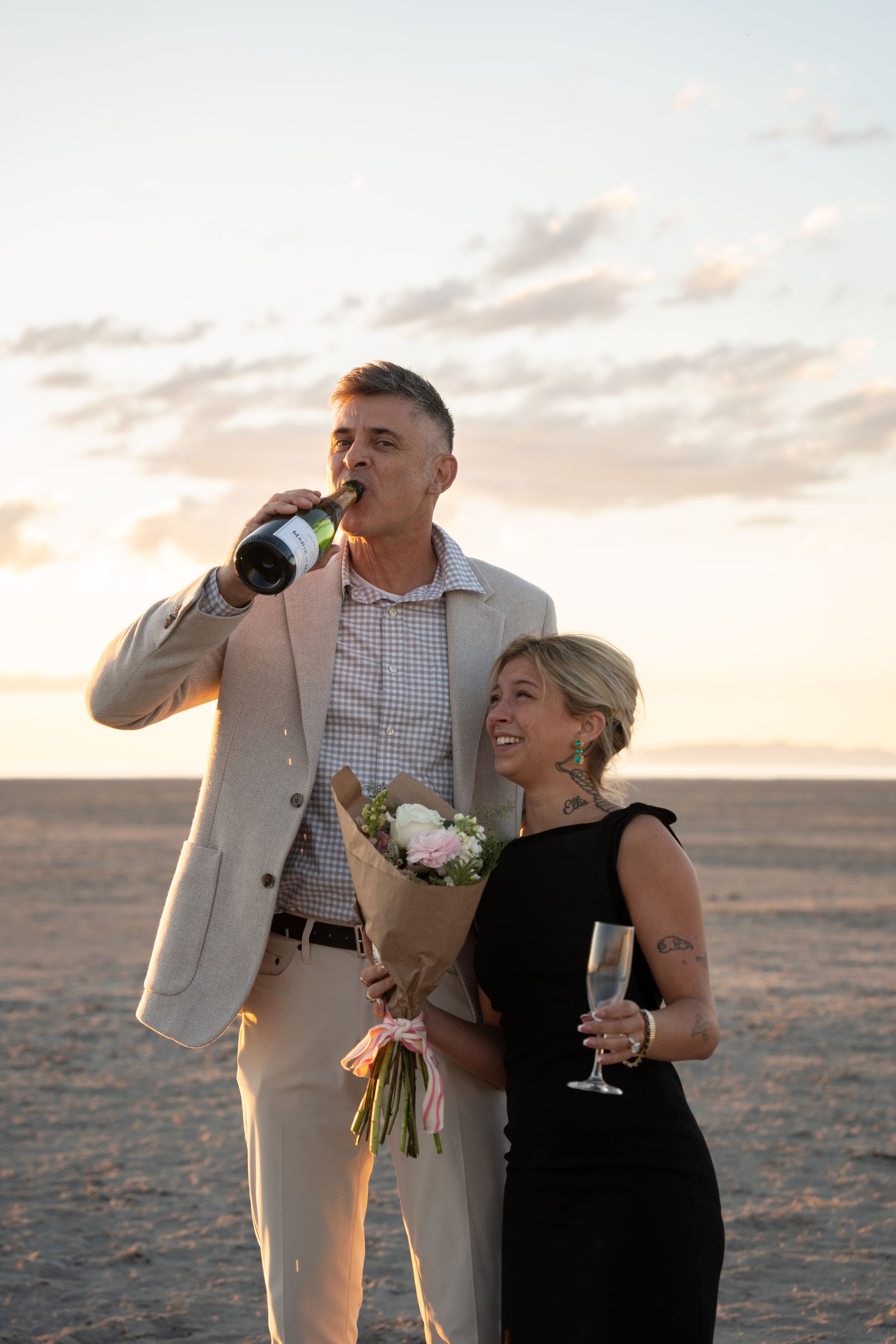 engaged Couple celebrating at sunset on salt flats, man drinking champagne from bottle, woman in black dress holding bouquet and wine glass.