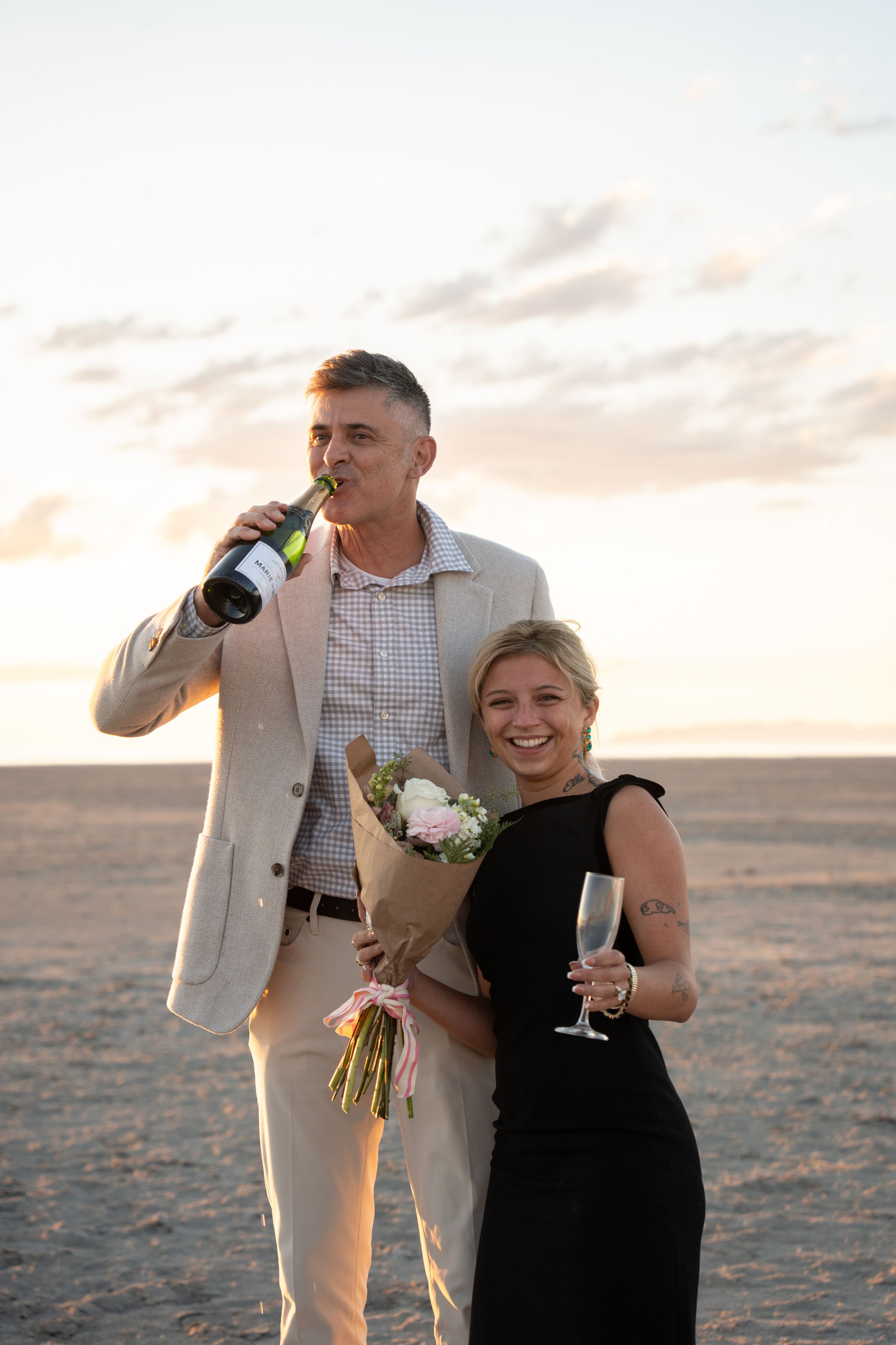 Couple celebrating proposal on salt flats at sunset, man drinking champagne bottle, woman in black dress holding flowers, champagne glass.