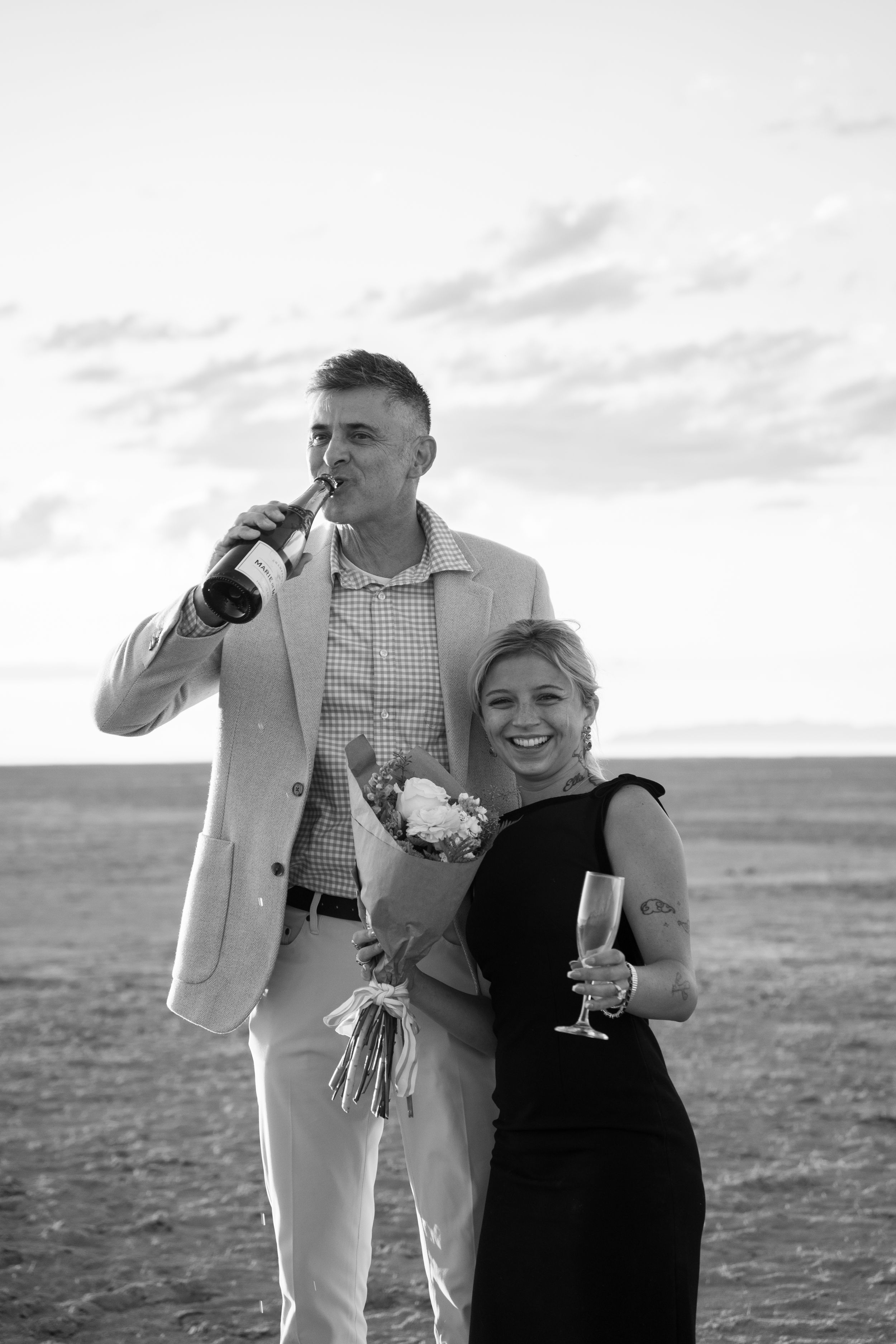 Black and white photo of a couple celebrating proposal, man drinking from bottle, woman in black dress holding champagne and flowers.