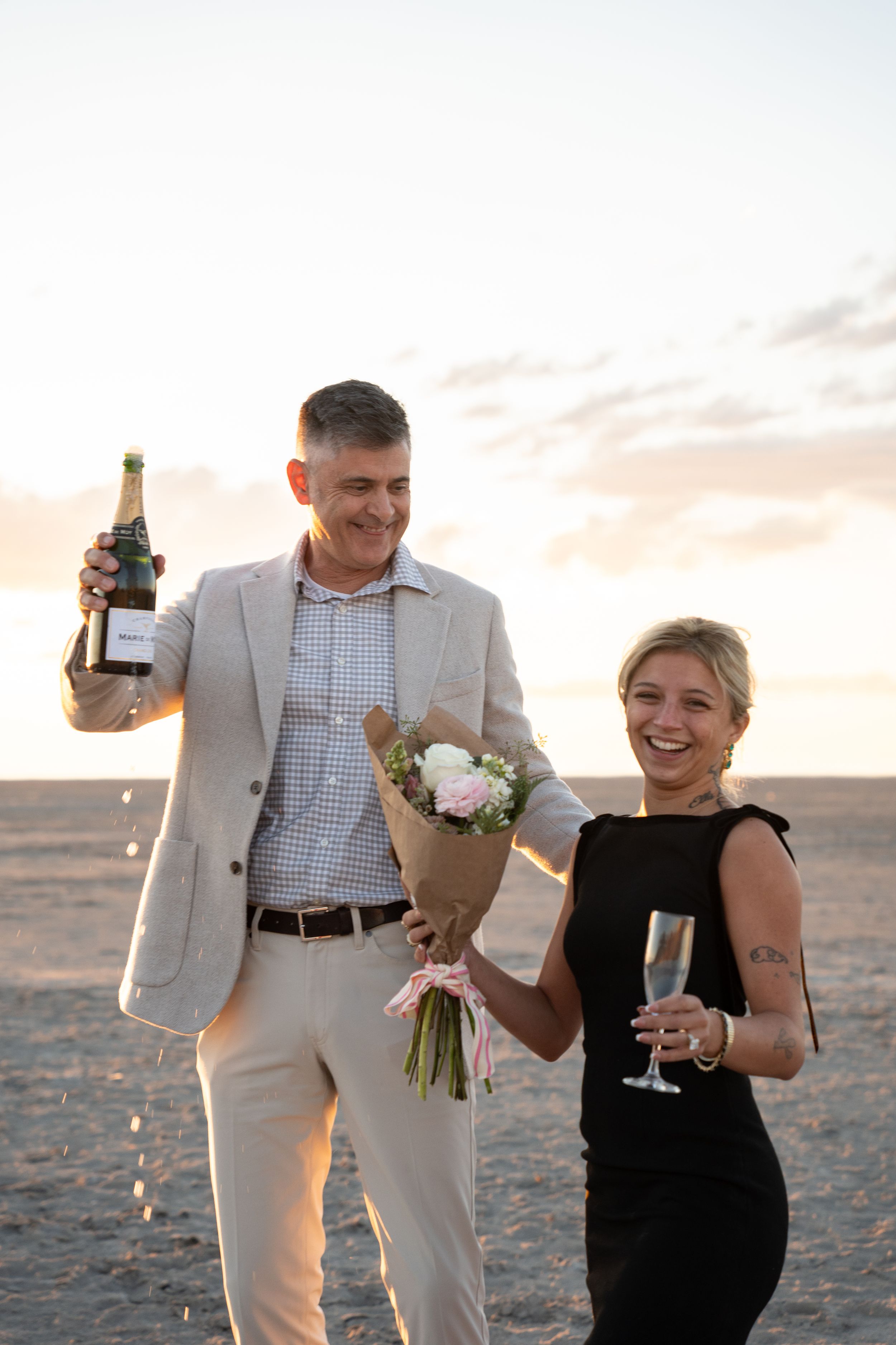 Couple celebrating on salt flats at sunset, man holding champagne bottle and flowers, woman in black dress laughing with champagne glass.