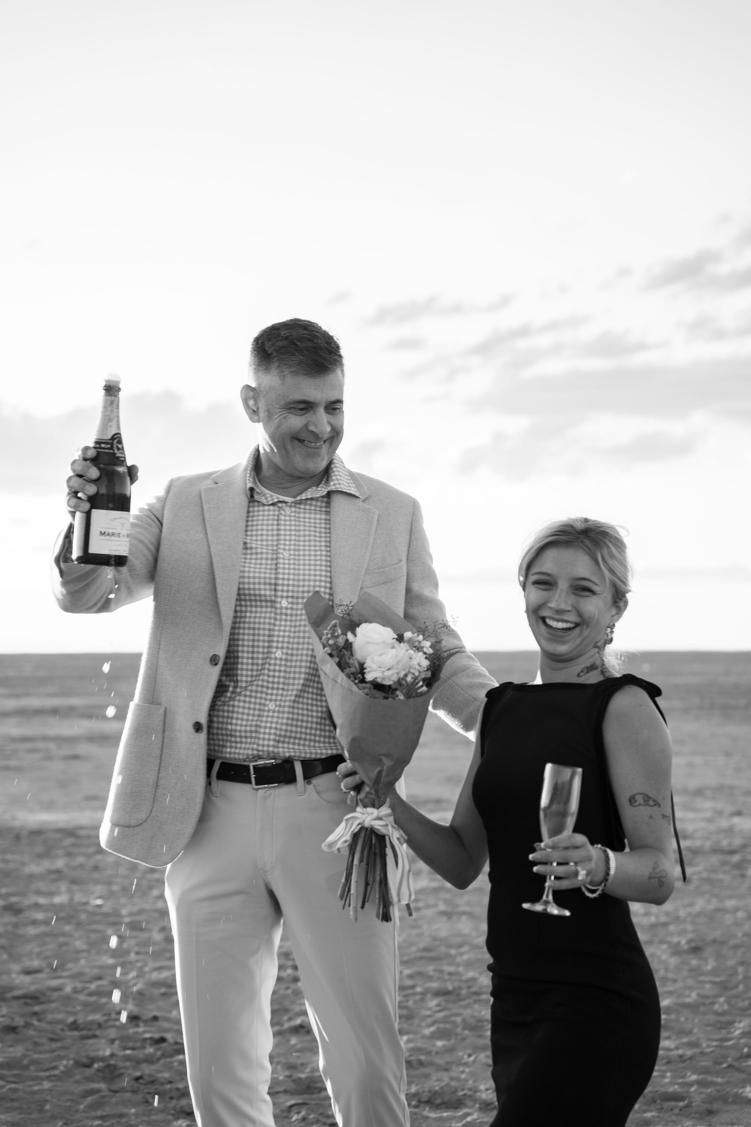Black and white photo of a couple celebrating on a salt flats, man holding champagne bottle and flowers, woman smiling with a glass.