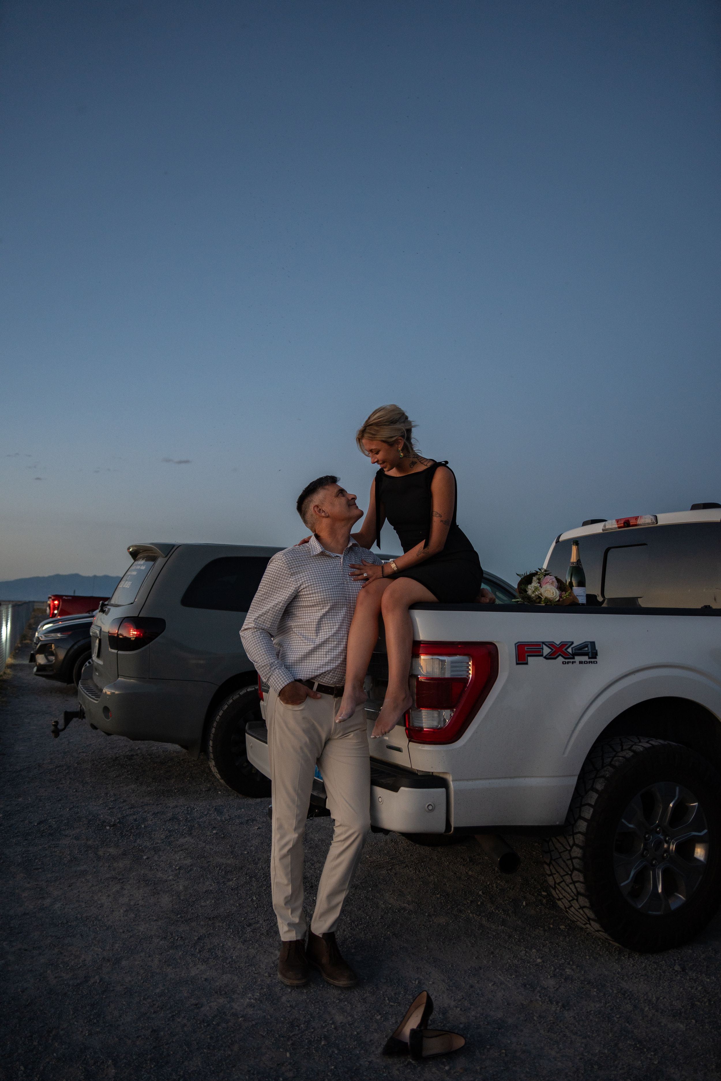 A couple shares a romantic moment at dusk, woman sitting on white Ford F-150 tailgate while man stands close.