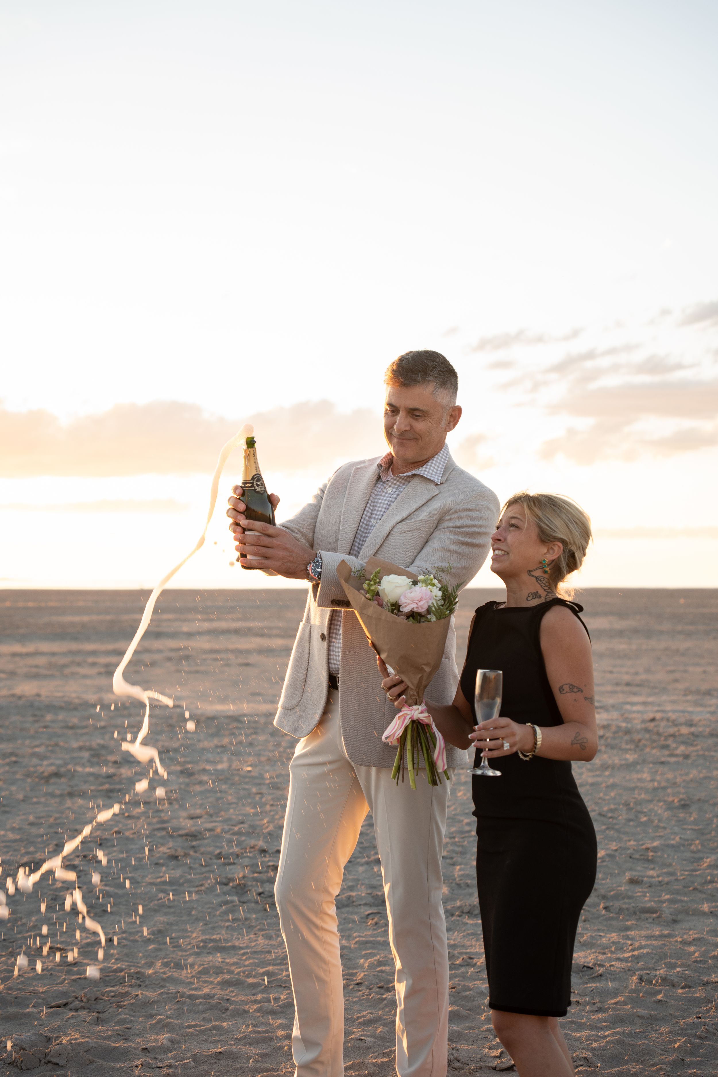 Couple celebrating proposal on salt lake at sunset, man popping champagne bottle, woman in black dress holding flowers and champagne glass.