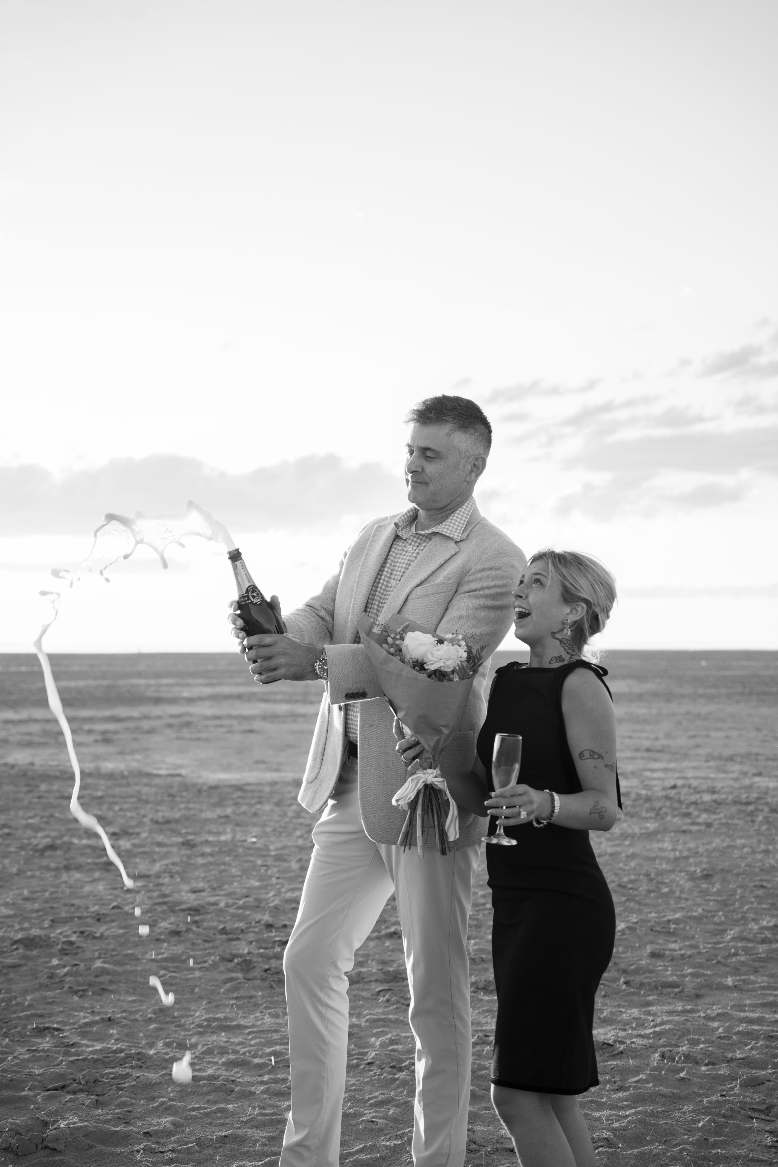 Black and white photo of couple celebrating on flat backdrop, man popping champagne bottle while woman holds a glass.