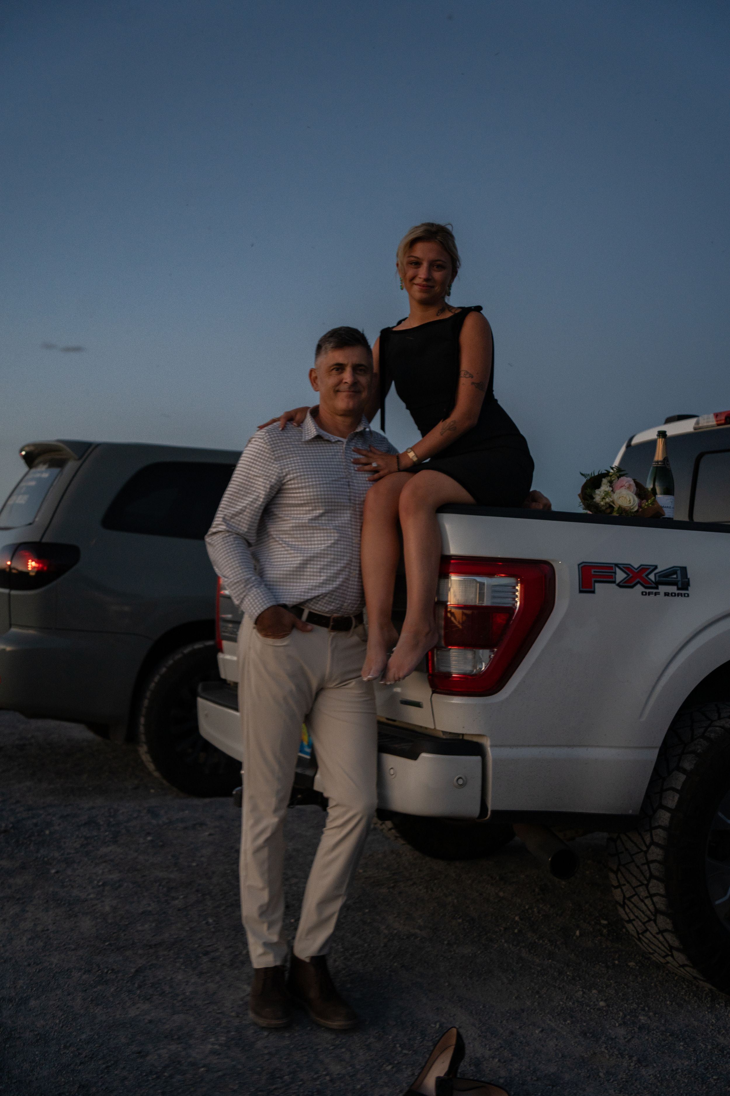 Couple posing at dusk by a white Ford F-150 FX4 pickup truck, man in white pants and woman in black dress.