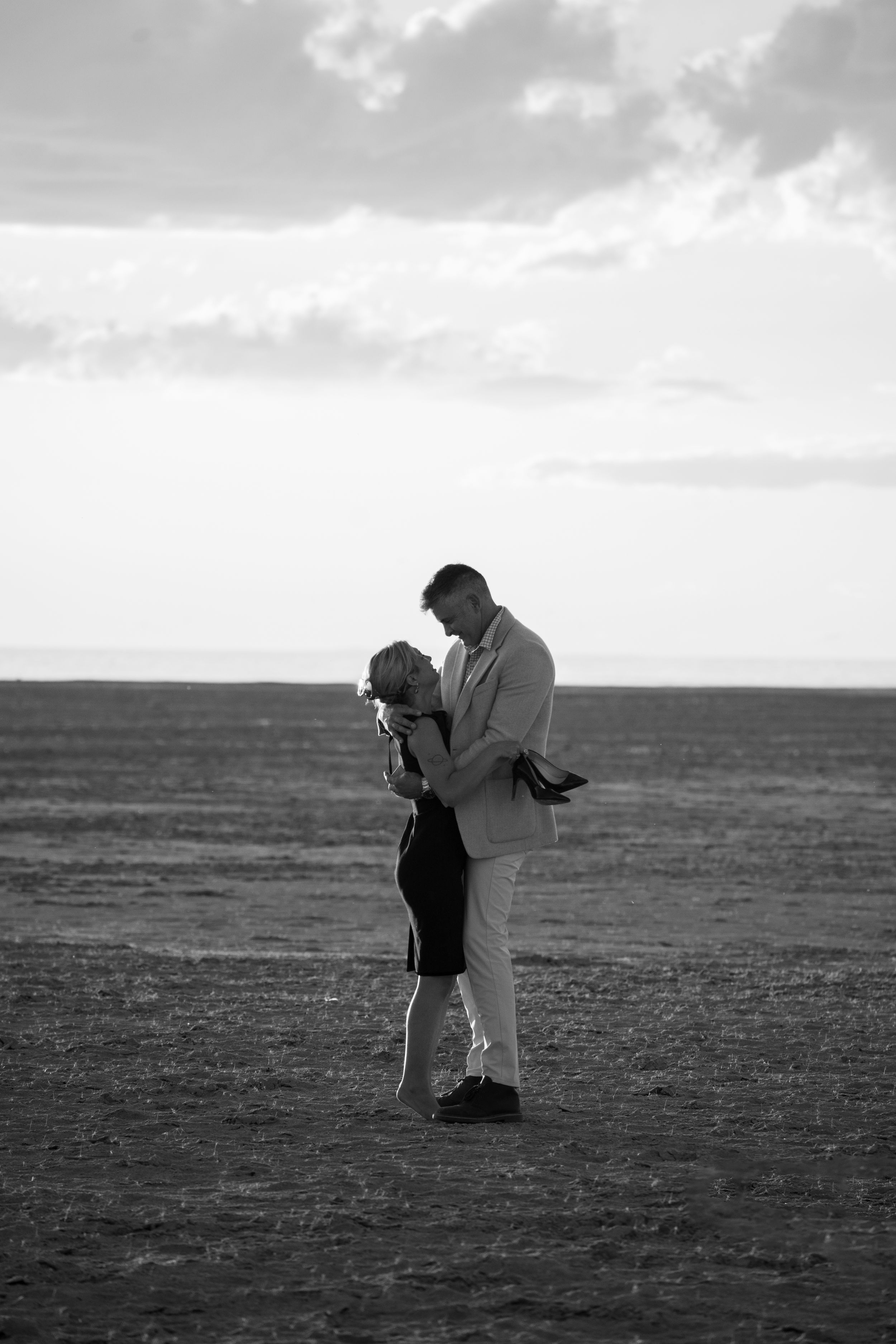 Black and white photo of a romantic couple embracing in an open field under a cloudy sky.