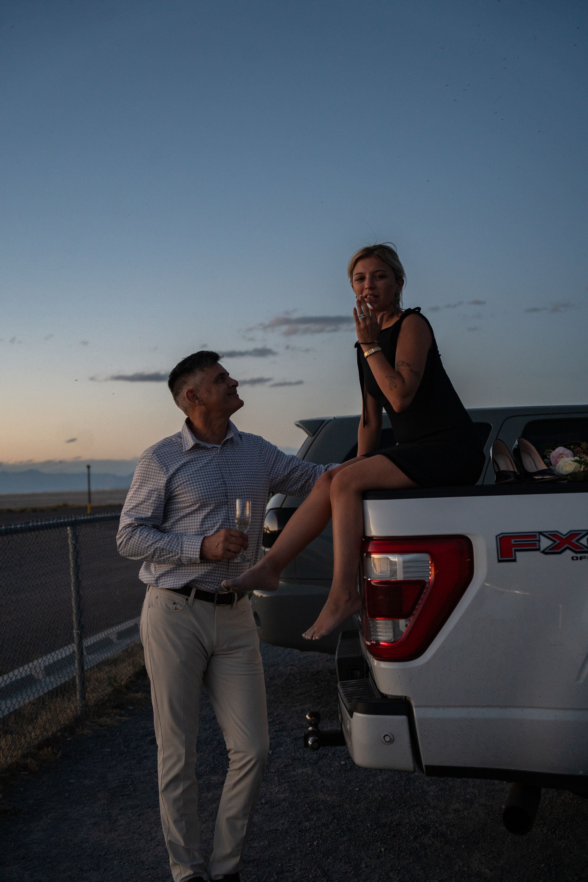 Couple sharing champagne by a Ford truck at sunset, woman sitting on tailgate in black dress, man in white shirt to celebrate proposal.