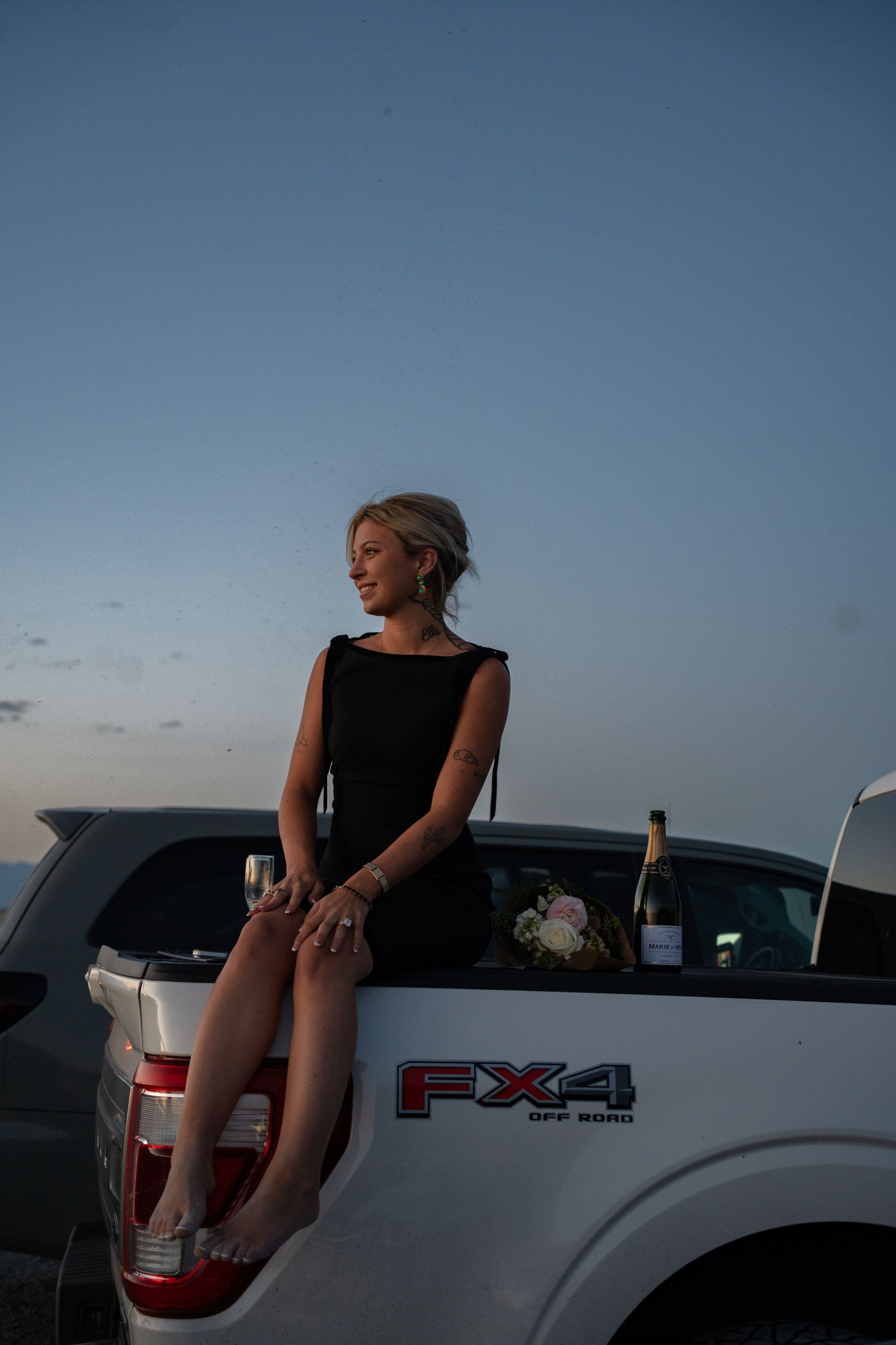 Woman in black dress sitting on Ford FX4 truck bed at dusk, holding champagne with flowers nearby.