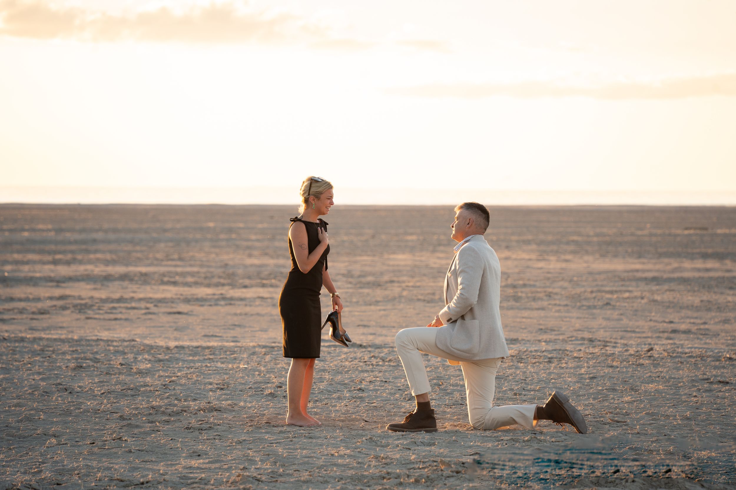 Man proposing on one knee to woman in black dress at sunset on a vast desert landscape at salt flats