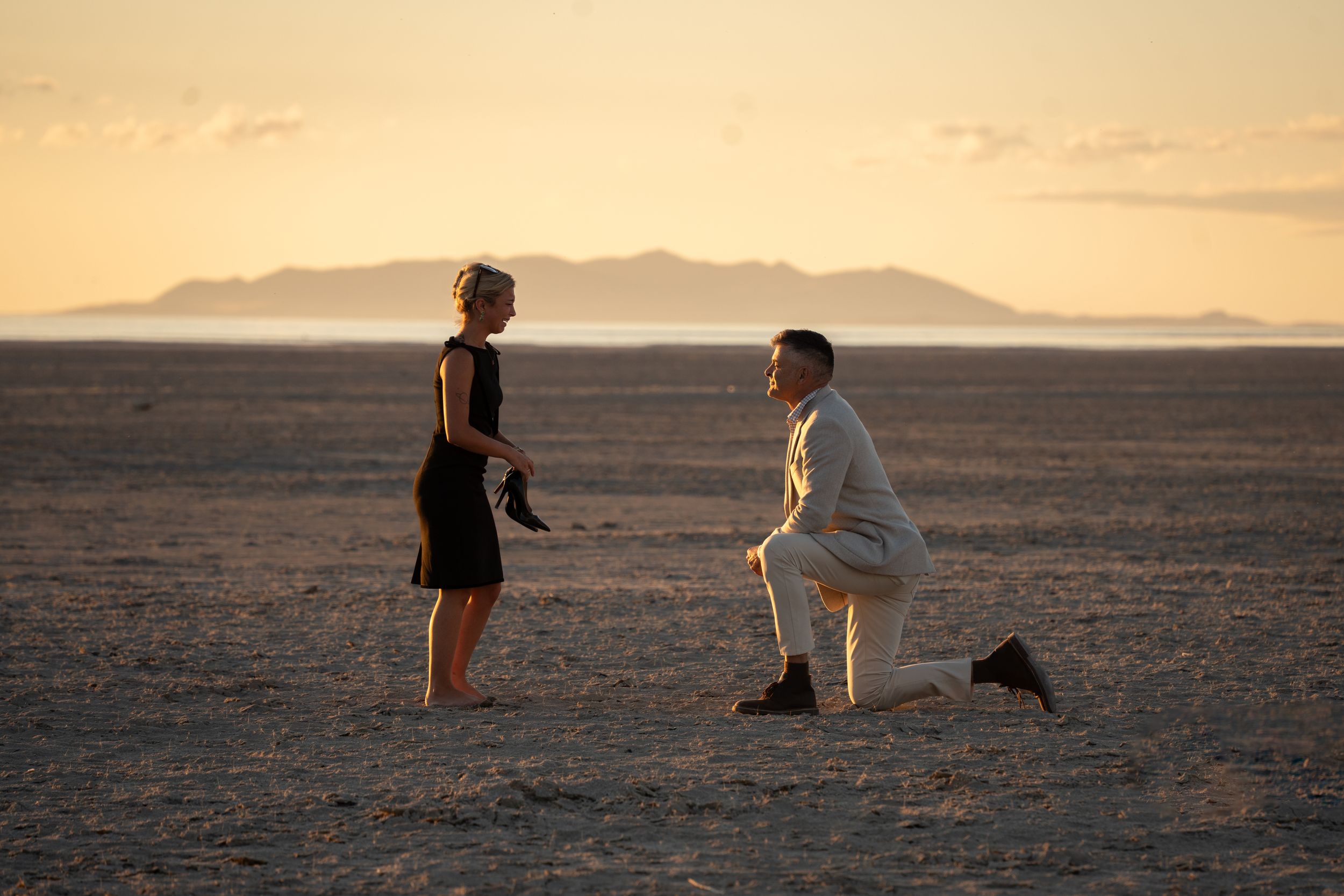 Man proposing on one knee to woman in black dress on vast desert salt flat at golden sunset.