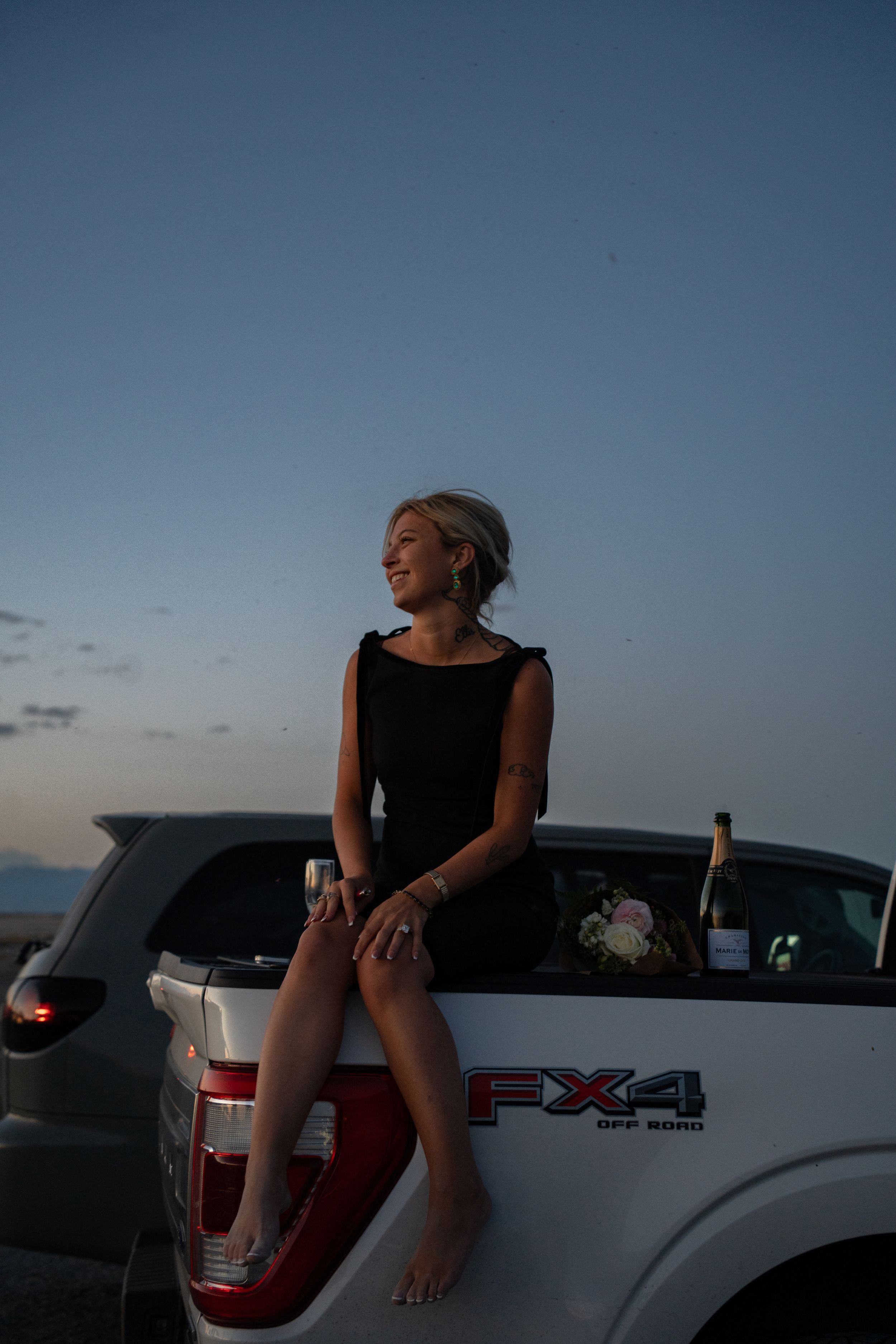 Woman in black dress sitting on tailgate of white Ford FX4 truck at dusk, smiling with champagne nearby.