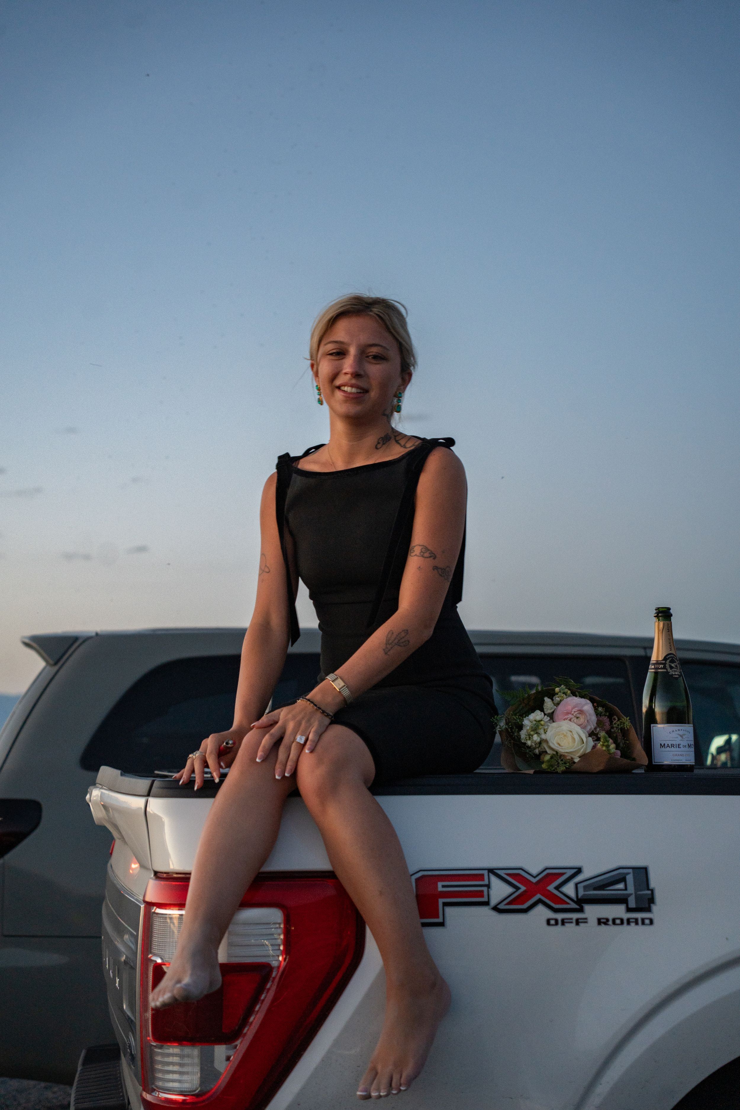 Woman in black dress sitting on Ford FX4 Off Road pickup truck tailgate at dusk with champagne and flowers.