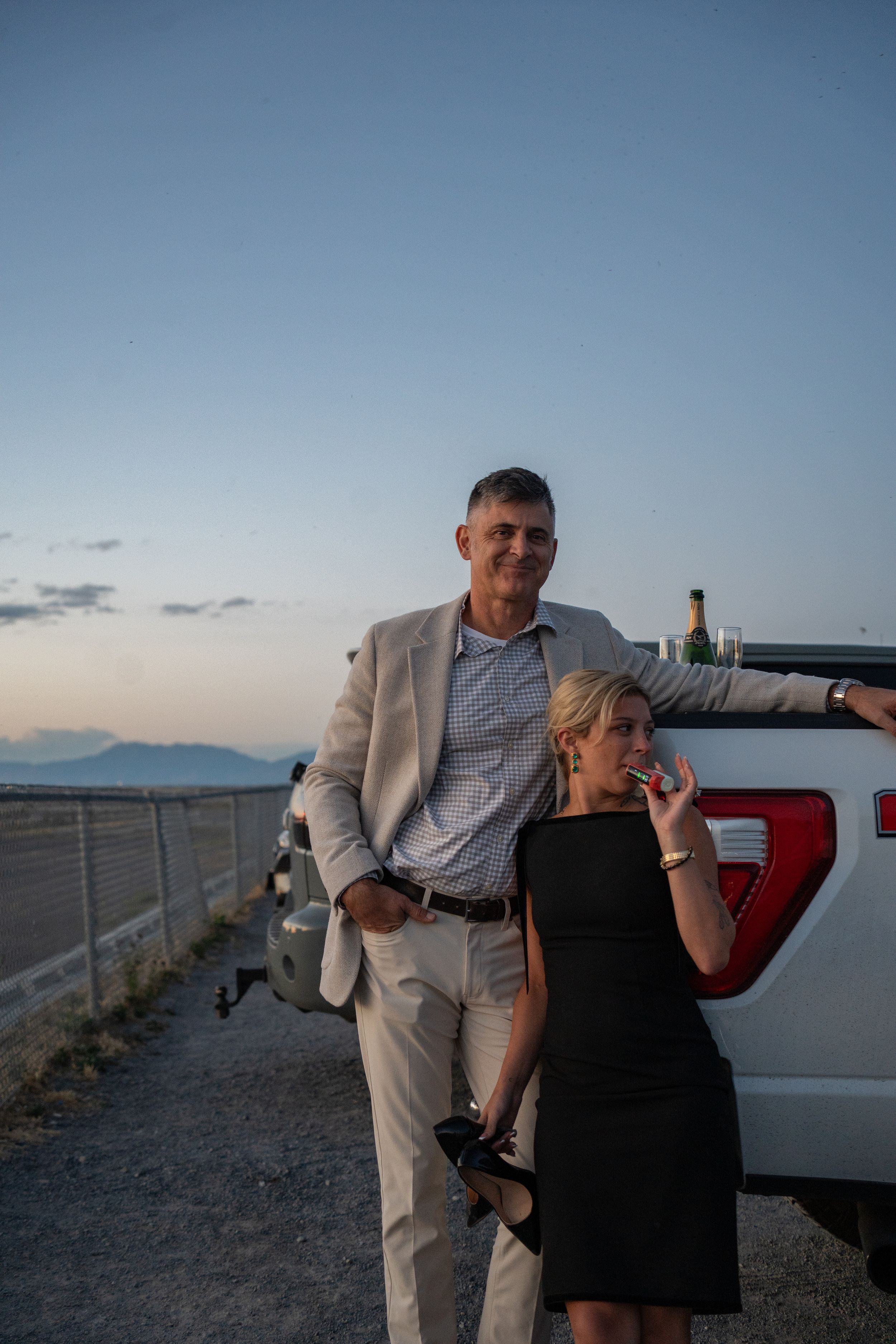 Couple posing by a white pickup truck at sunset, man in beige suit, woman in black dress holding a drink to celebrate their proposal