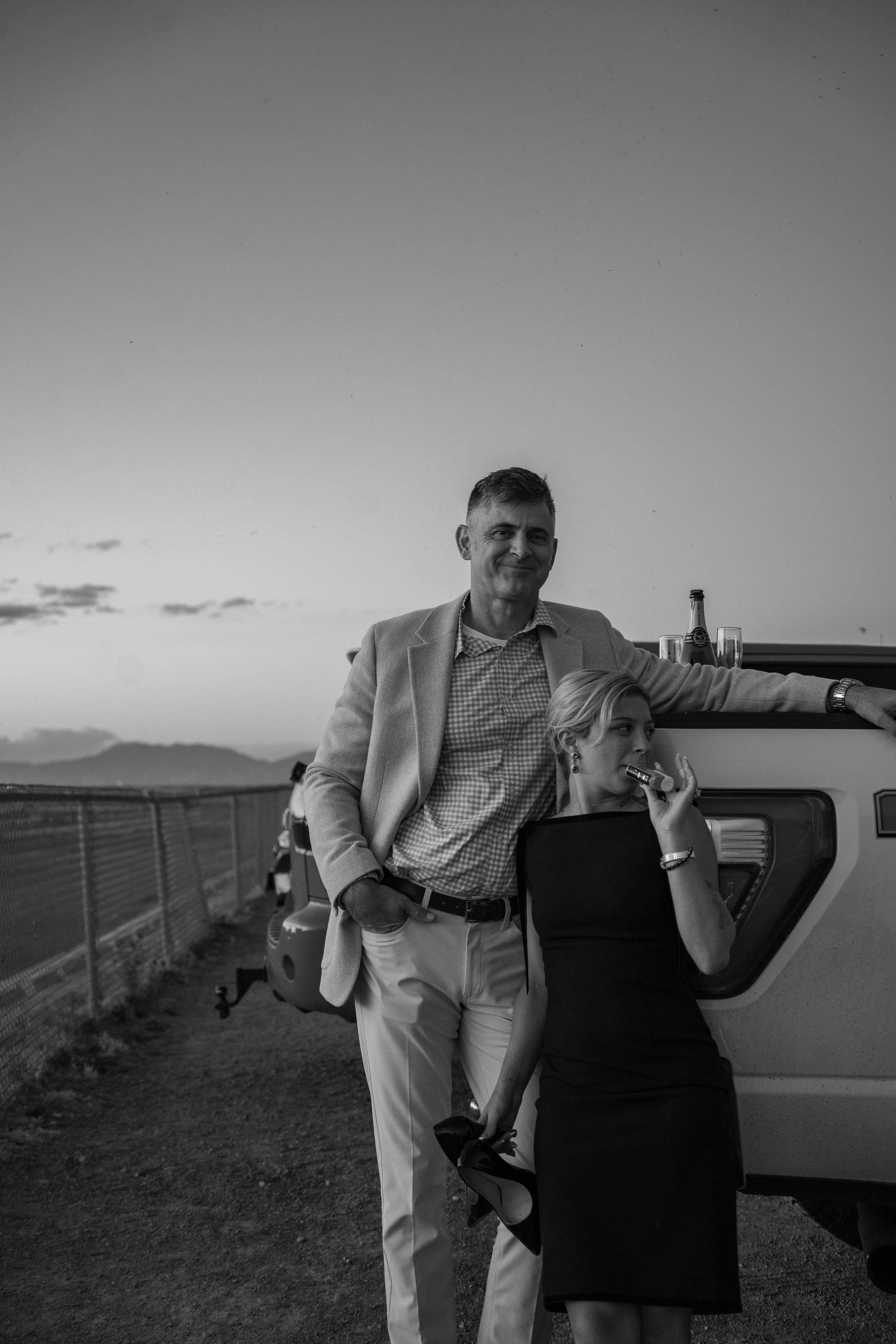 elegant Black and white photo of a couple leaning against a vehicle at sunset, man in blazer, woman in dress holding a glass.