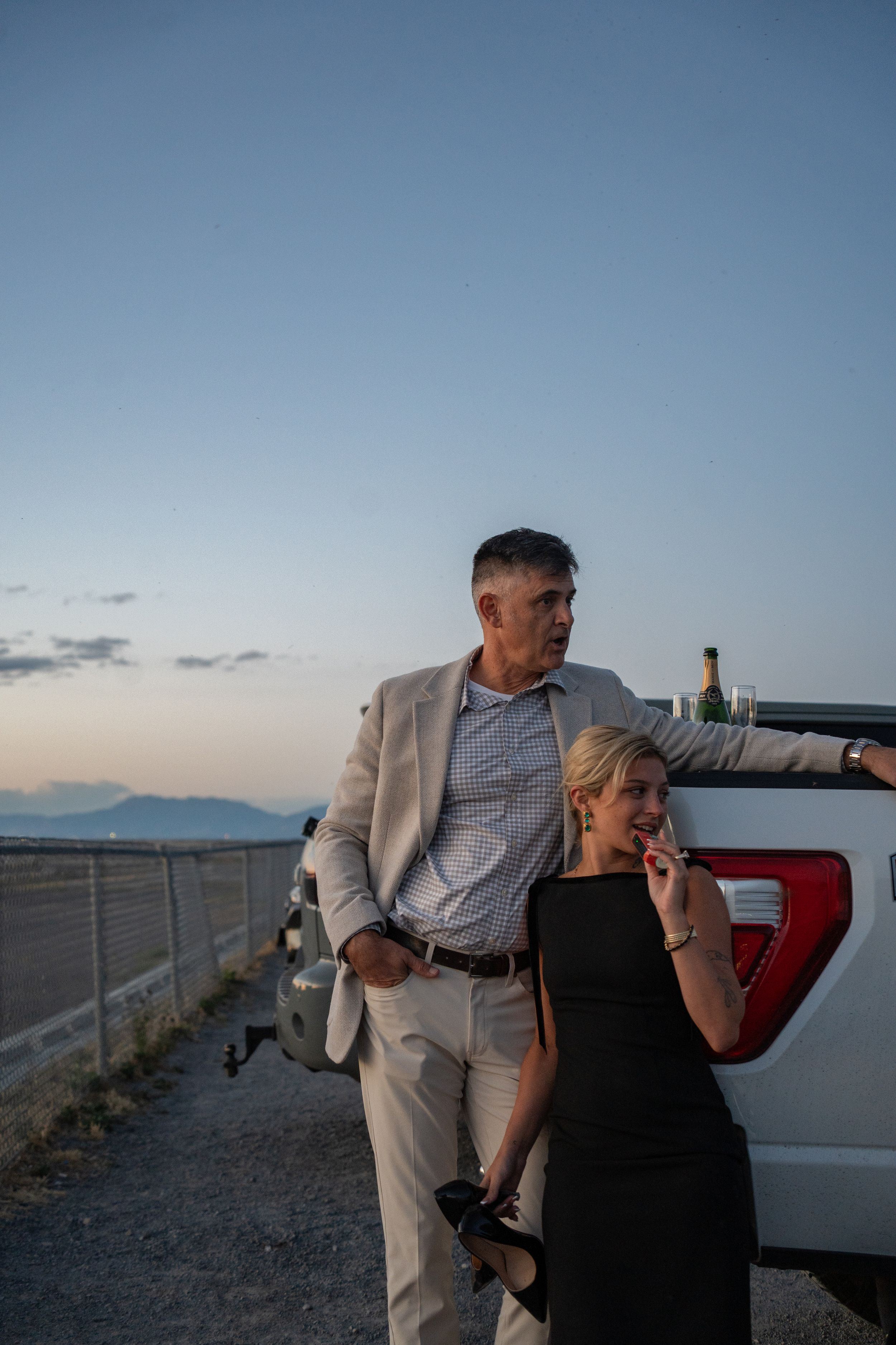 Couple in elegant attire leaning against white pickup truck at dusk with wasatch mountain backdrop.