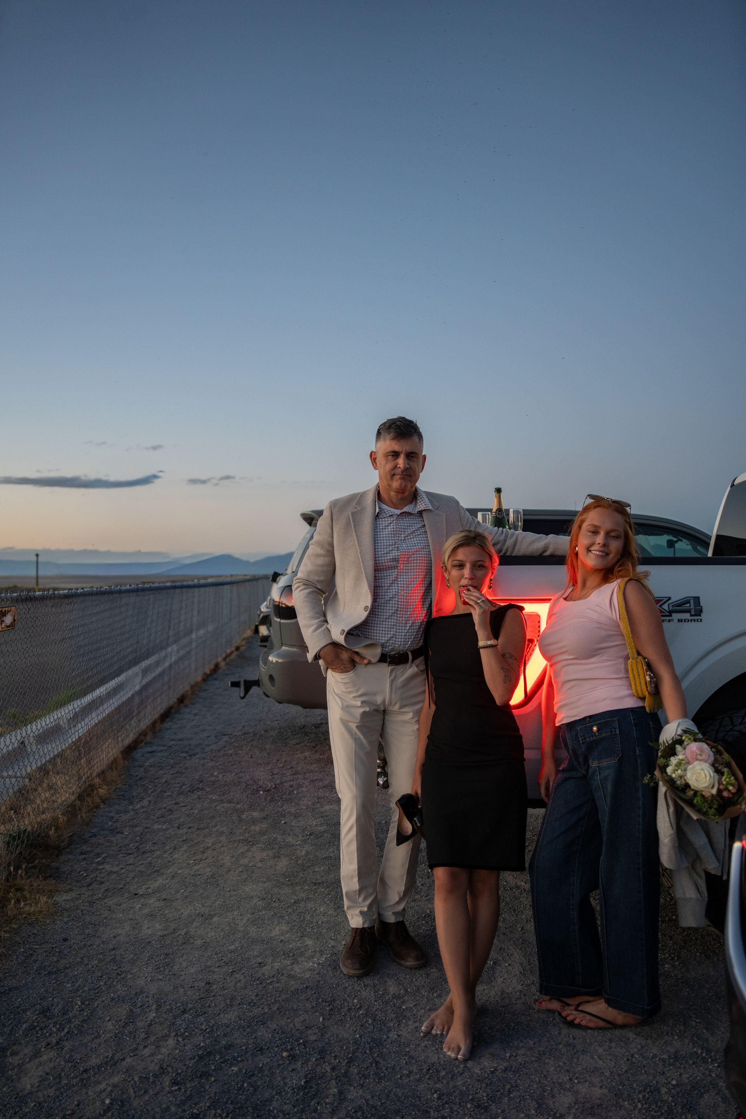 A man and his girlfriend and best friend pose by a truck at dusk near a waterfront, dressed in semi-formal attire celebrating a proposal