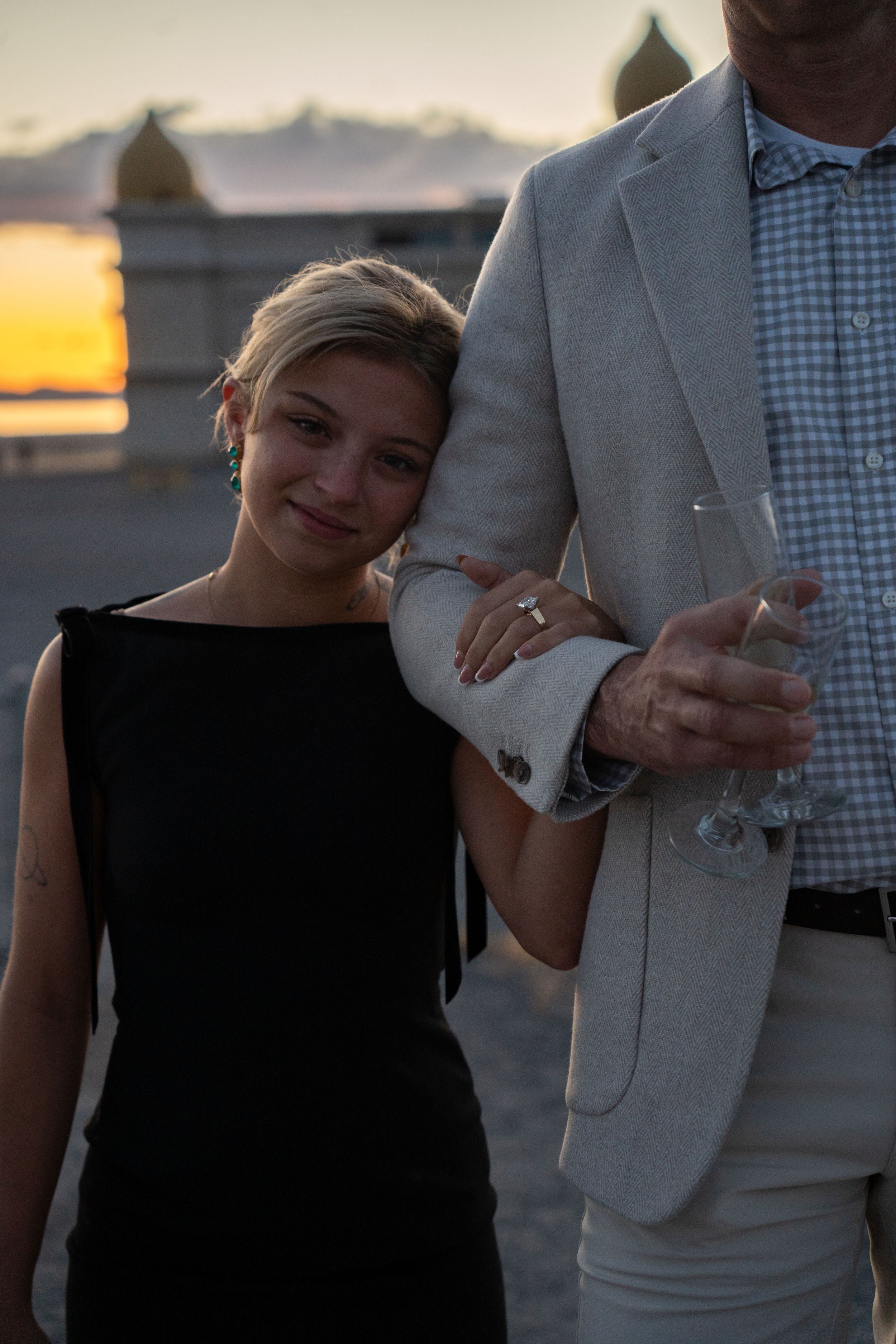 A young woman in a black dress and engagement ring leans on a man in a light suit holding champagne at sunset.