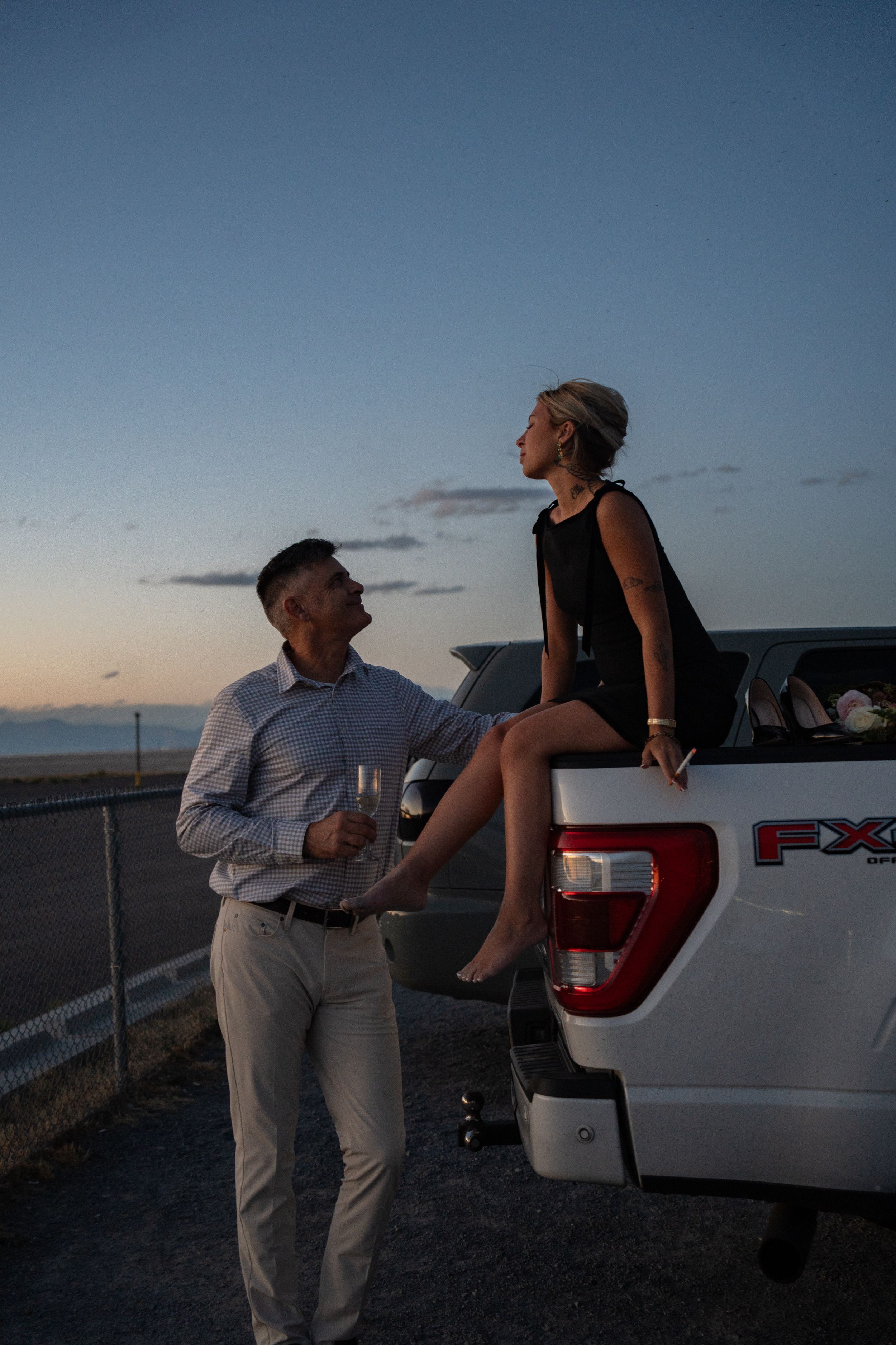 Couple sharing a romantic sunset moment by a white Ford FX pickup truck, man holding champagne glass.