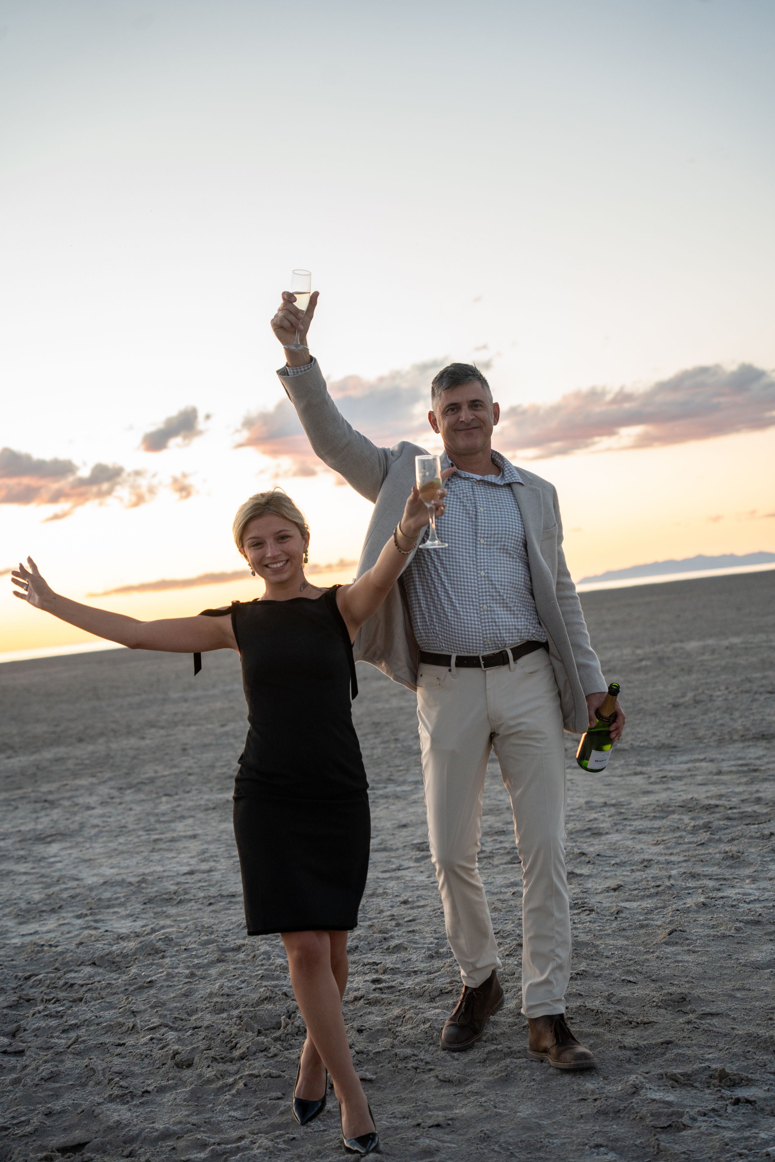 Couple celebrating on salt flats at sunset, raising champagne glasses, woman in black dress, man in light suit holding champagne bottle