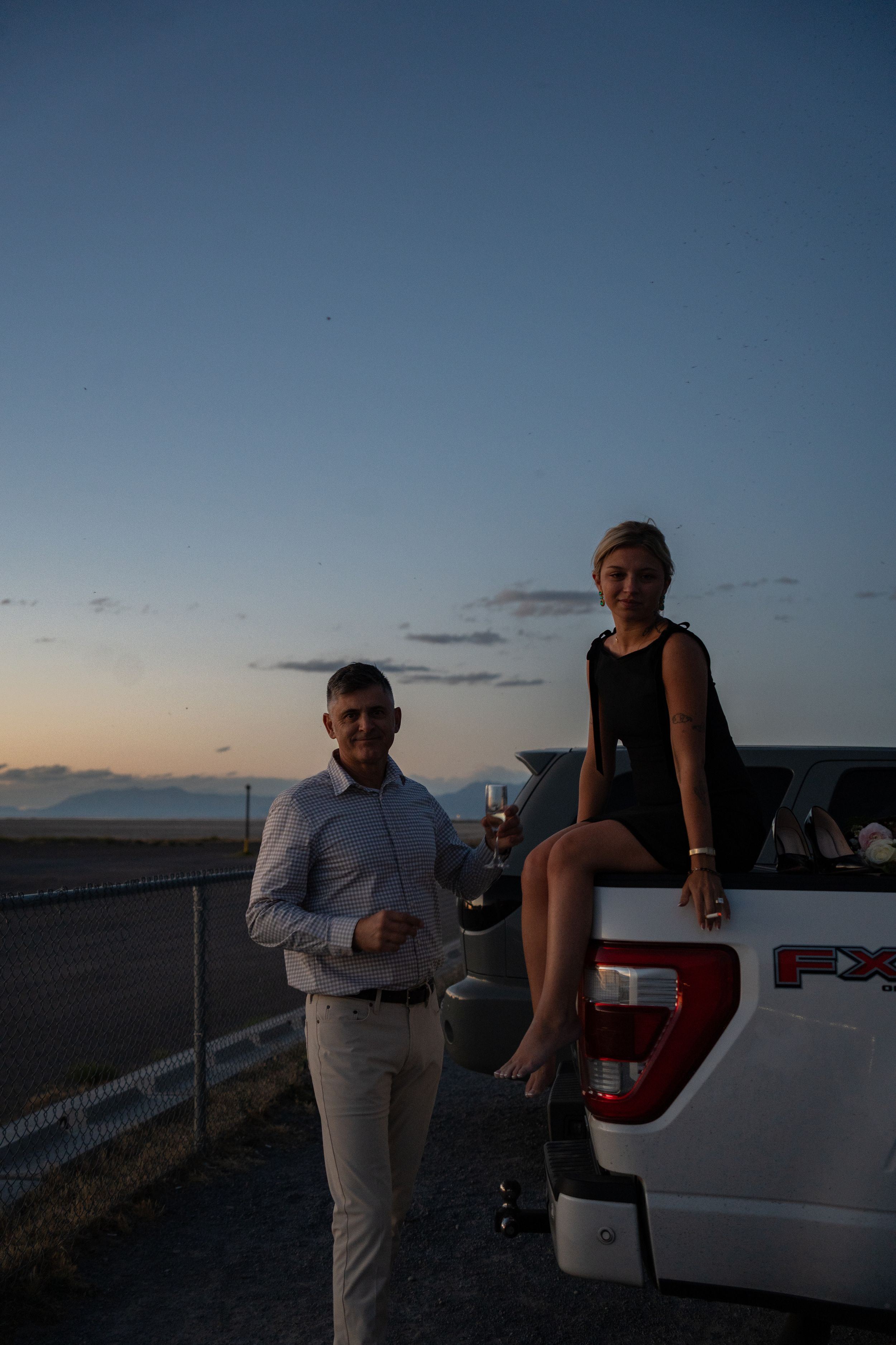 Couple celebrating at dusk beside a white Ford truck, woman sitting on tailgate holding champagne glass.