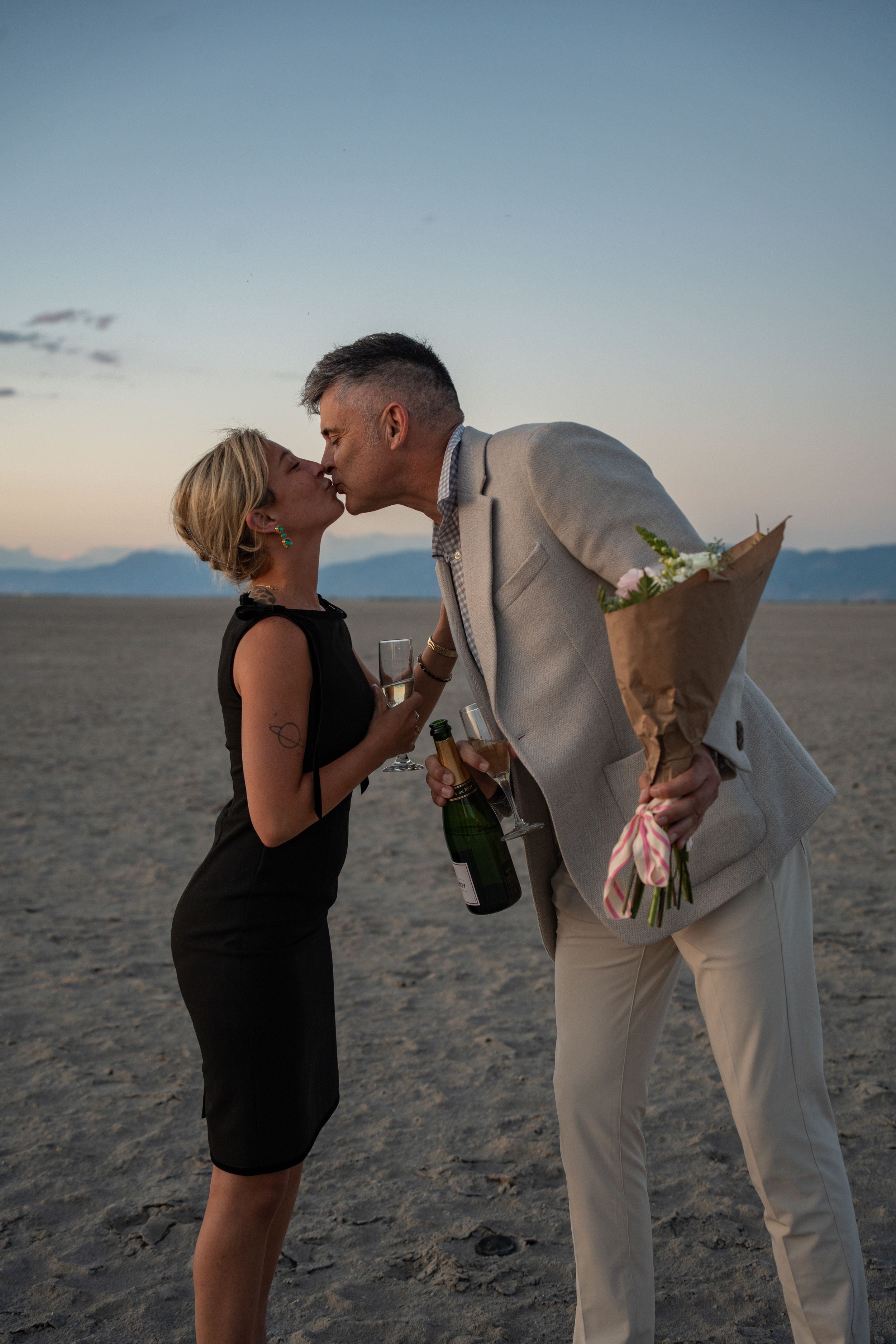 couple kissing while holding champagne and flowers to celebrate a proposal